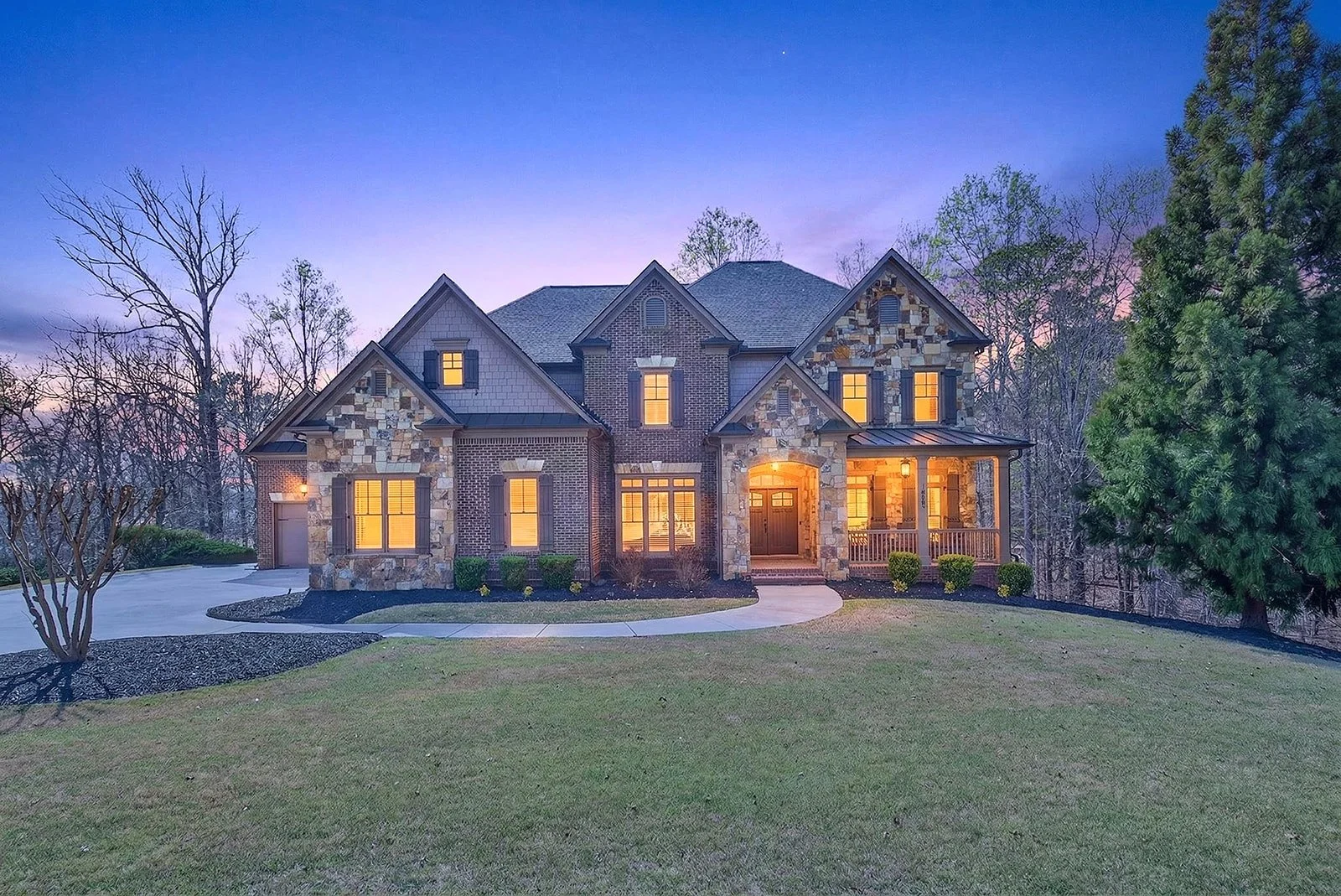 A large two-story house with illuminated windows, a mix of stone and brick exterior, a front porch, surrounded by a lawn and trees at dusk.
