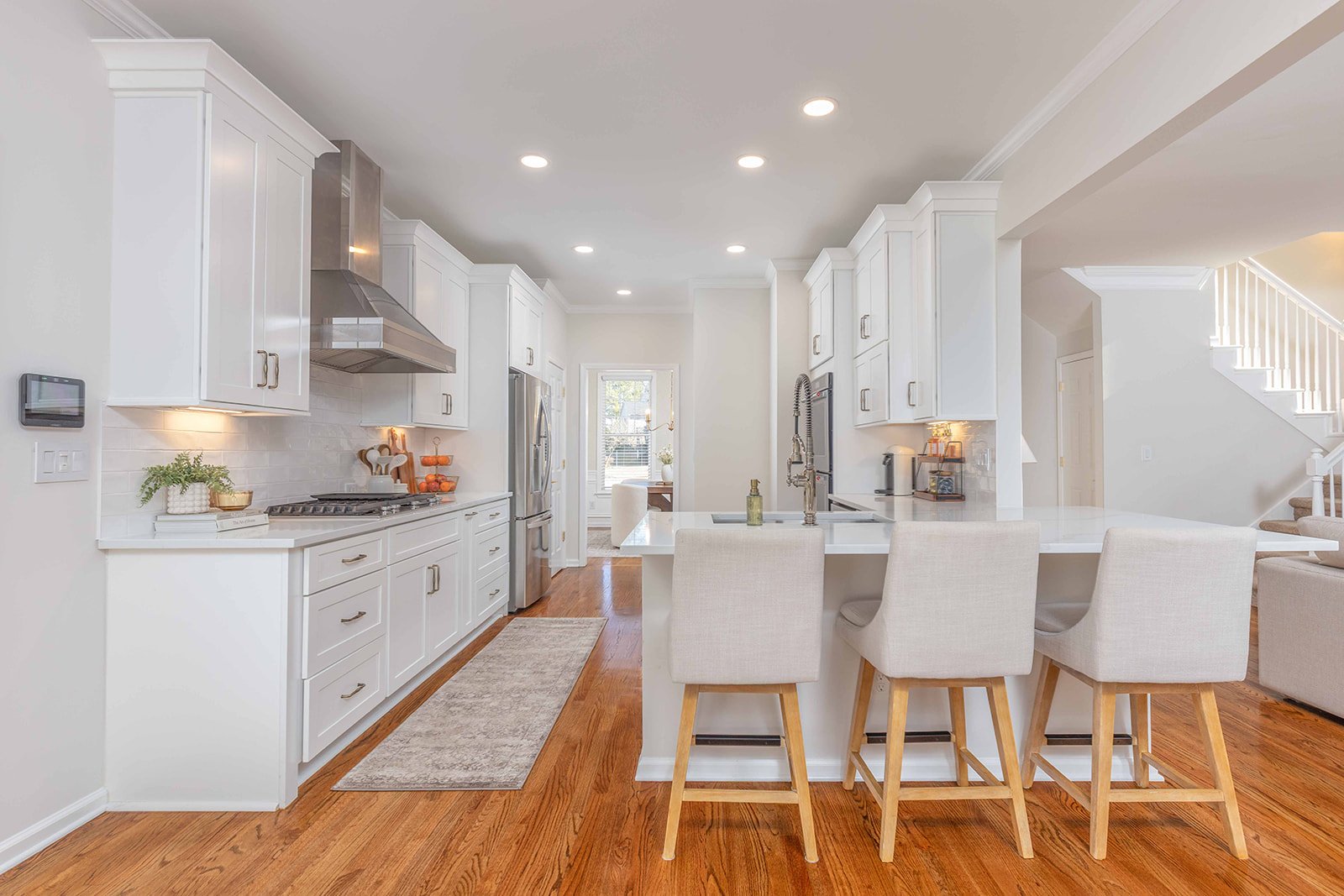 A bright, modern kitchen with white cabinets, a kitchen island with beige chairs, stainless steel appliances, hardwood floors, and decorative accents.