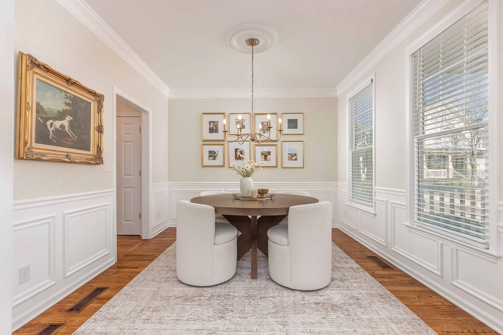 A dining room with a round wooden table, four white upholstered chairs, a chandelier, framed photographs on the wall, large windows with white blinds, a beige rug, and a vase with flowers on the table.