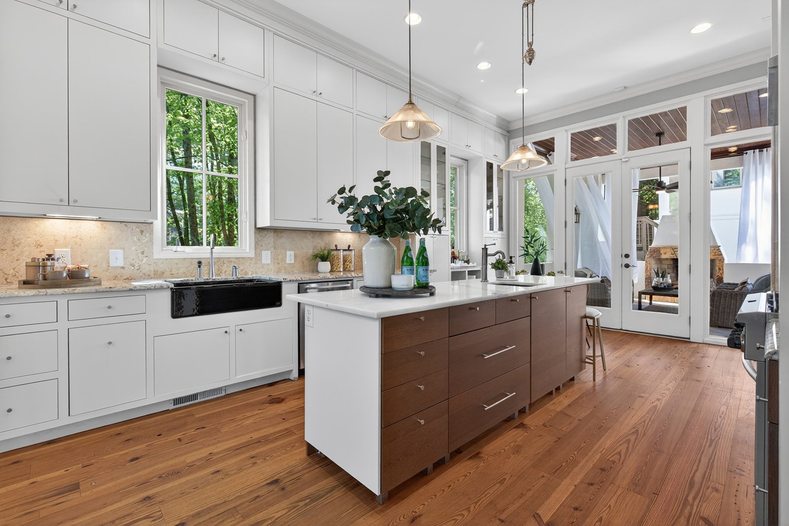 Modern kitchen with white cabinets, a large island with wooden drawers, and natural light coming through windows and glass doors. Hardwood floors and pendant lighting are also visible.