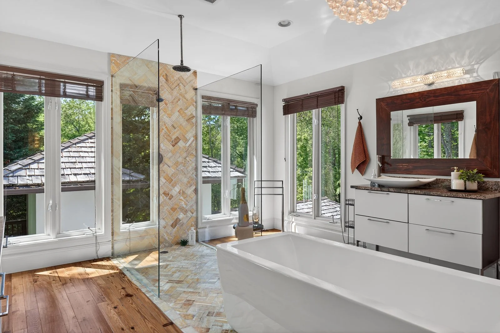 Modern bathroom with large windowed shower area, freestanding bathtub, white vanity with vessel sink, wooden framed mirror, and greenery outside.