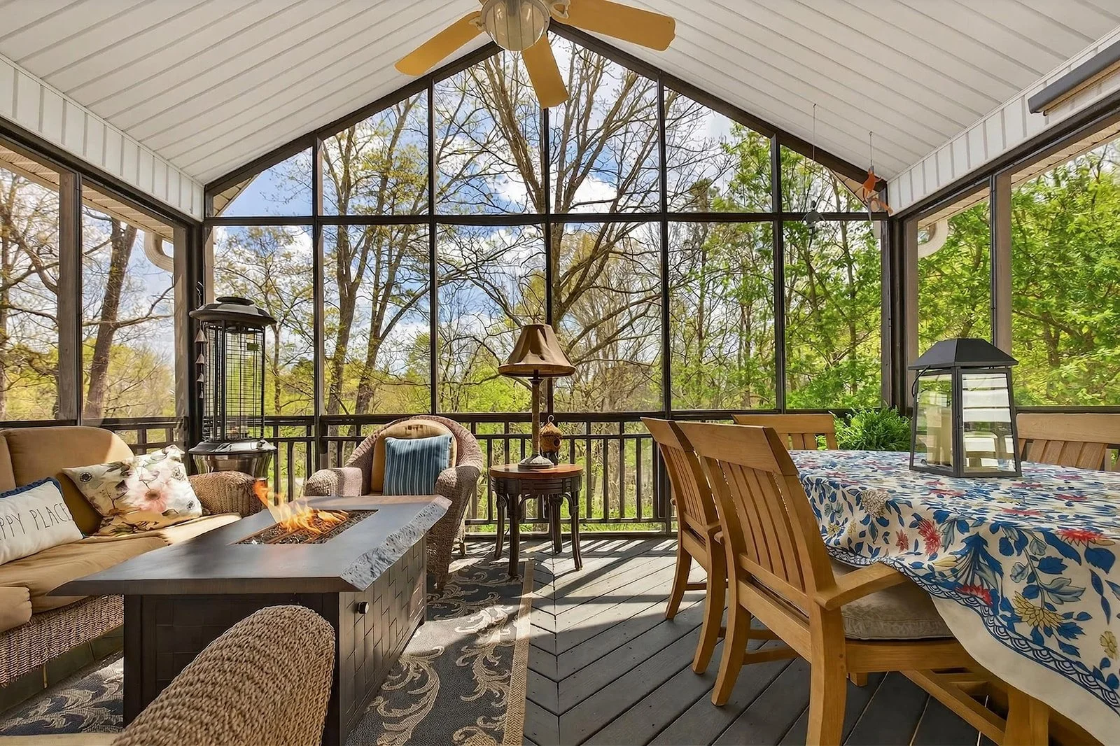 Sunroom with glass walls and ceiling, outdoor views, wicker and wooden furniture, a fire feature, and potted plants.