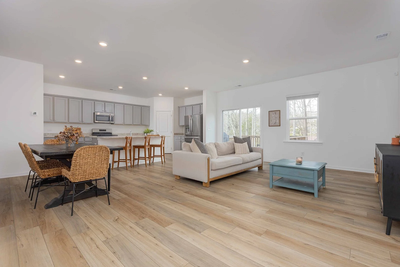 Open-concept living room and kitchen with light-colored walls, hardwood flooring, gray kitchen cabinets, stainless steel appliances, and a cream sofa. Dining area has a dark table with wicker chairs, and there is a blue coffee table near the sofa, wi