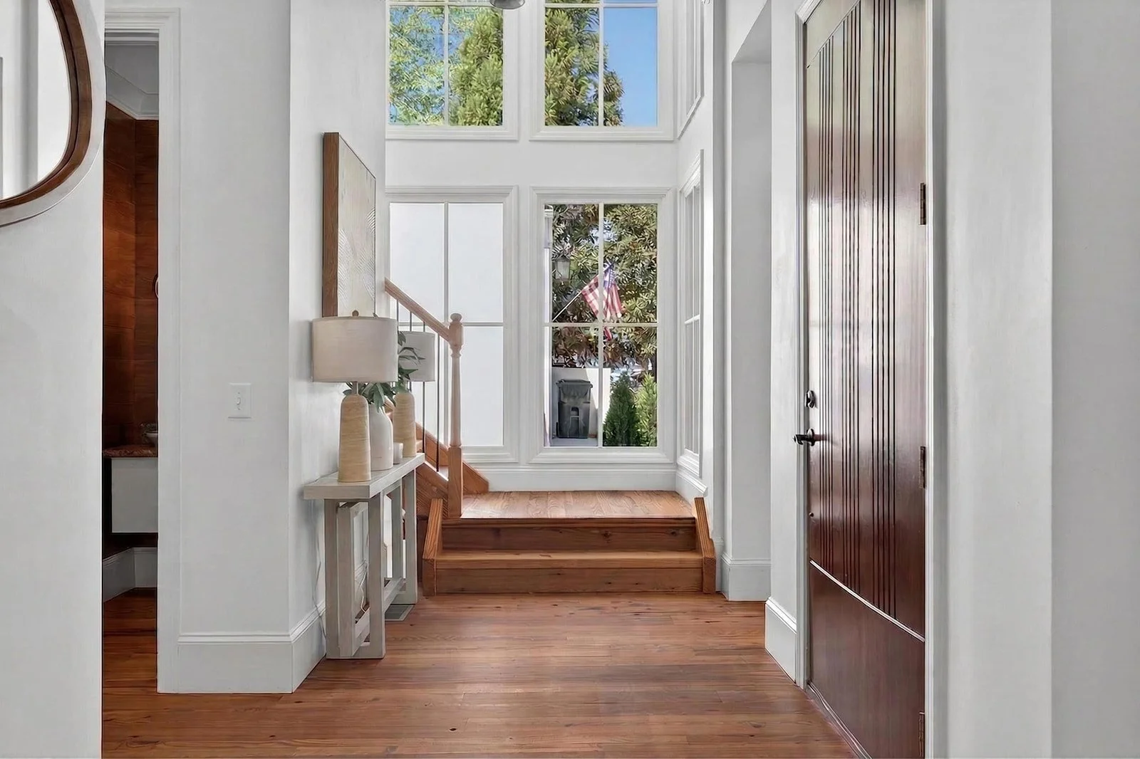 Entryway of a house with wooden flooring, a white console table with vases and a lamp, a staircase with wooden steps, large windows letting in natural light, and a brown wooden door.