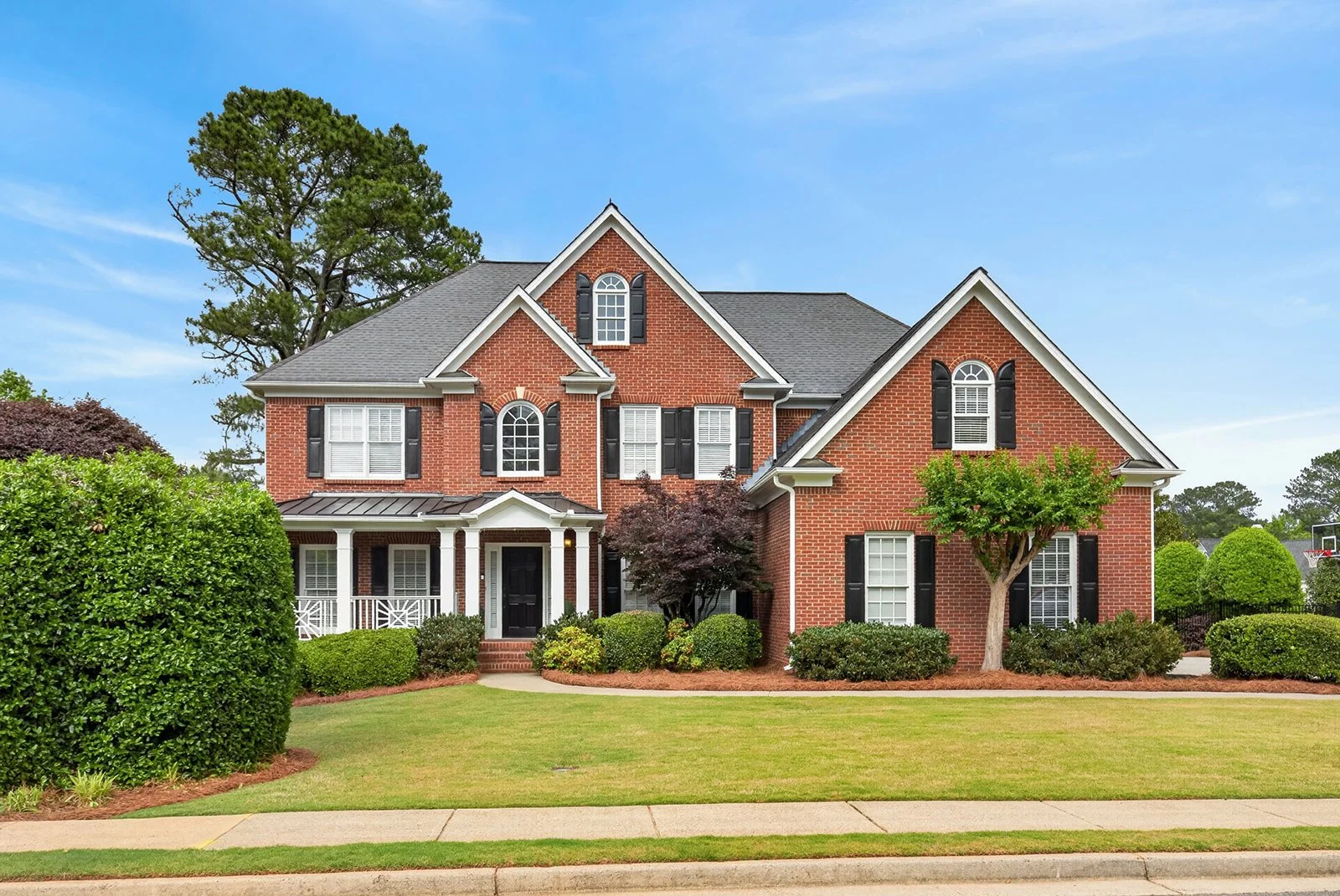 A large two-story brick house with white trim, black shutters, and a covered front porch, surrounded by a well-manicured lawn and various trees and shrubs, with a clear blue sky above.