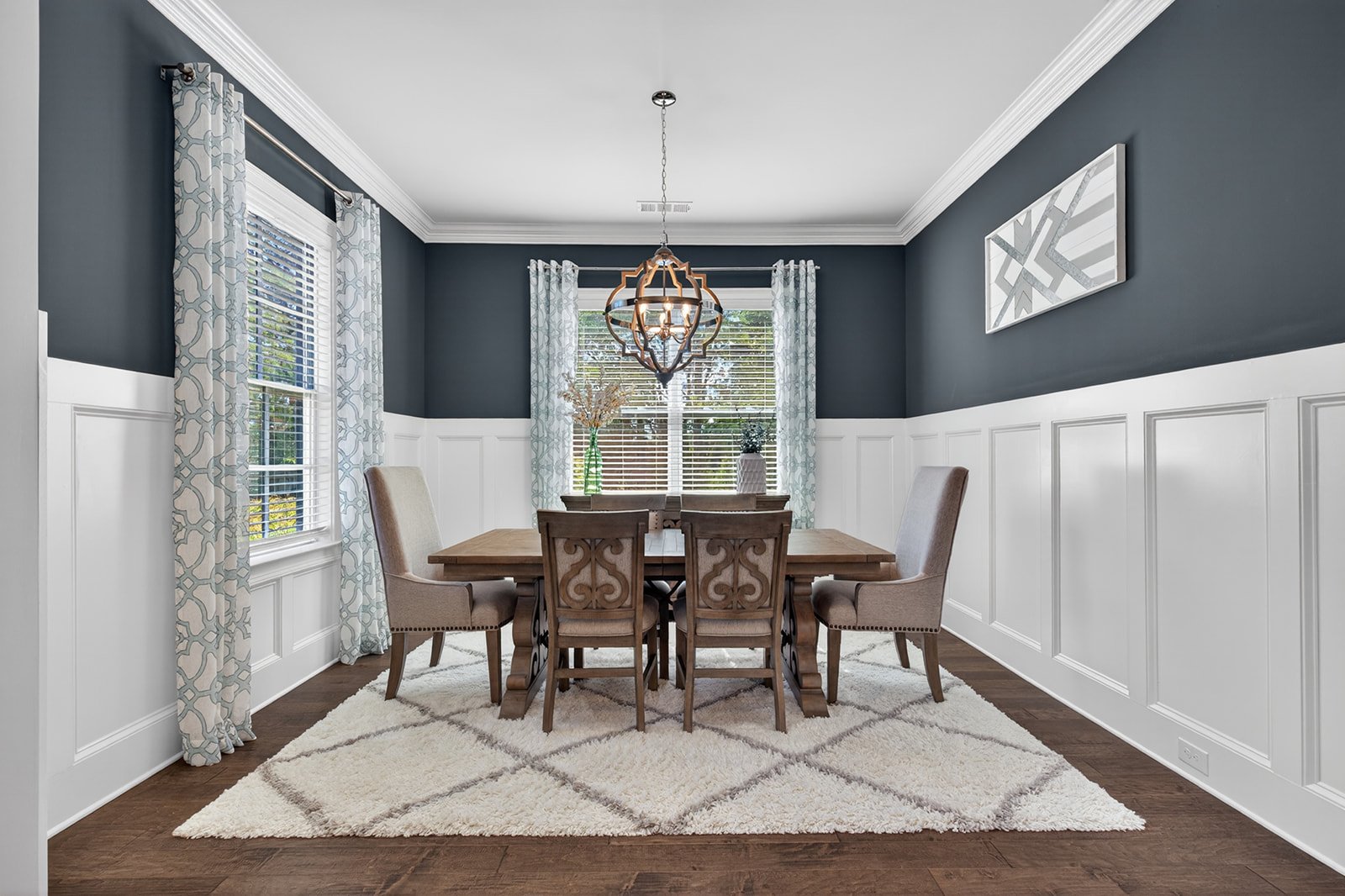 A dining room with dark blue upper walls and white wainscoting lower walls, featuring four windows with patterned curtains, a wooden dining table with six chairs, a beige rug, and a modern chandelier hanging above the table.