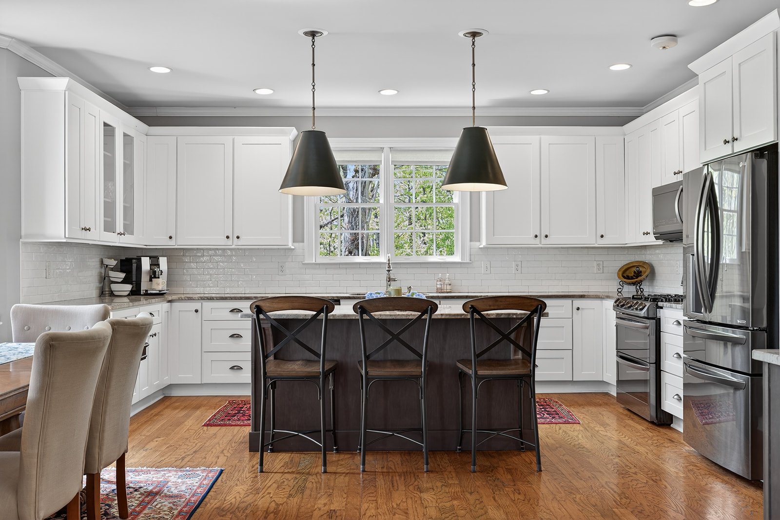 Modern kitchen with white cabinets, stainless steel appliances, black pendant lights, center island with three black chairs, and hardwood floors.