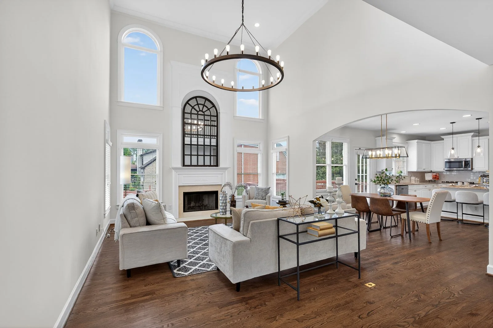 Open-concept living room and kitchen with white walls, large windows, a modern black chandelier, and hardwood floors, featuring cream sofas, a fireplace, and a dining table with chairs.