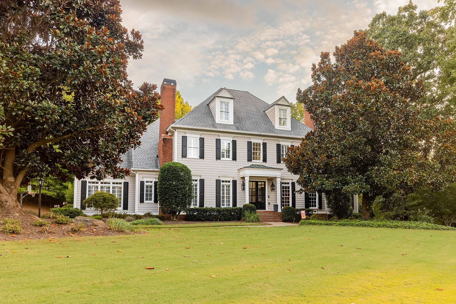 A large, white two-story house with black shutters, a front porch, and a brick chimney, surrounded by tall trees and a well-maintained lawn.