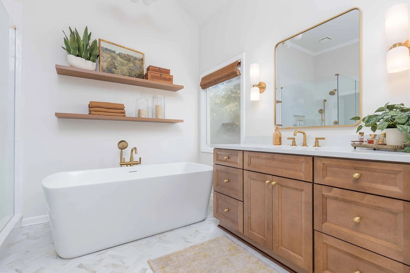 A modern bathroom with a white freestanding bathtub, wooden vanity with brass fixtures, mirror, two wall sconces, window with a bamboo shade, and floating shelves with decor, towels, and candles.
