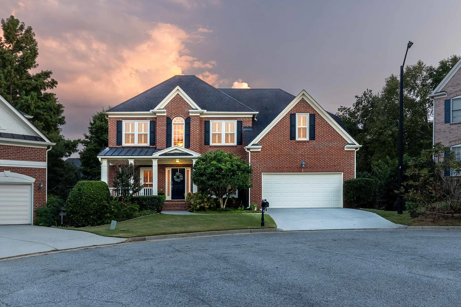 A two-story brick house with a black roof, white accents, and dark shutters. The front yard has neatly trimmed bushes, a small tree, and a manicured lawn. The driveway leads to a white garage door, and there is a mailbox on a black post near the curb