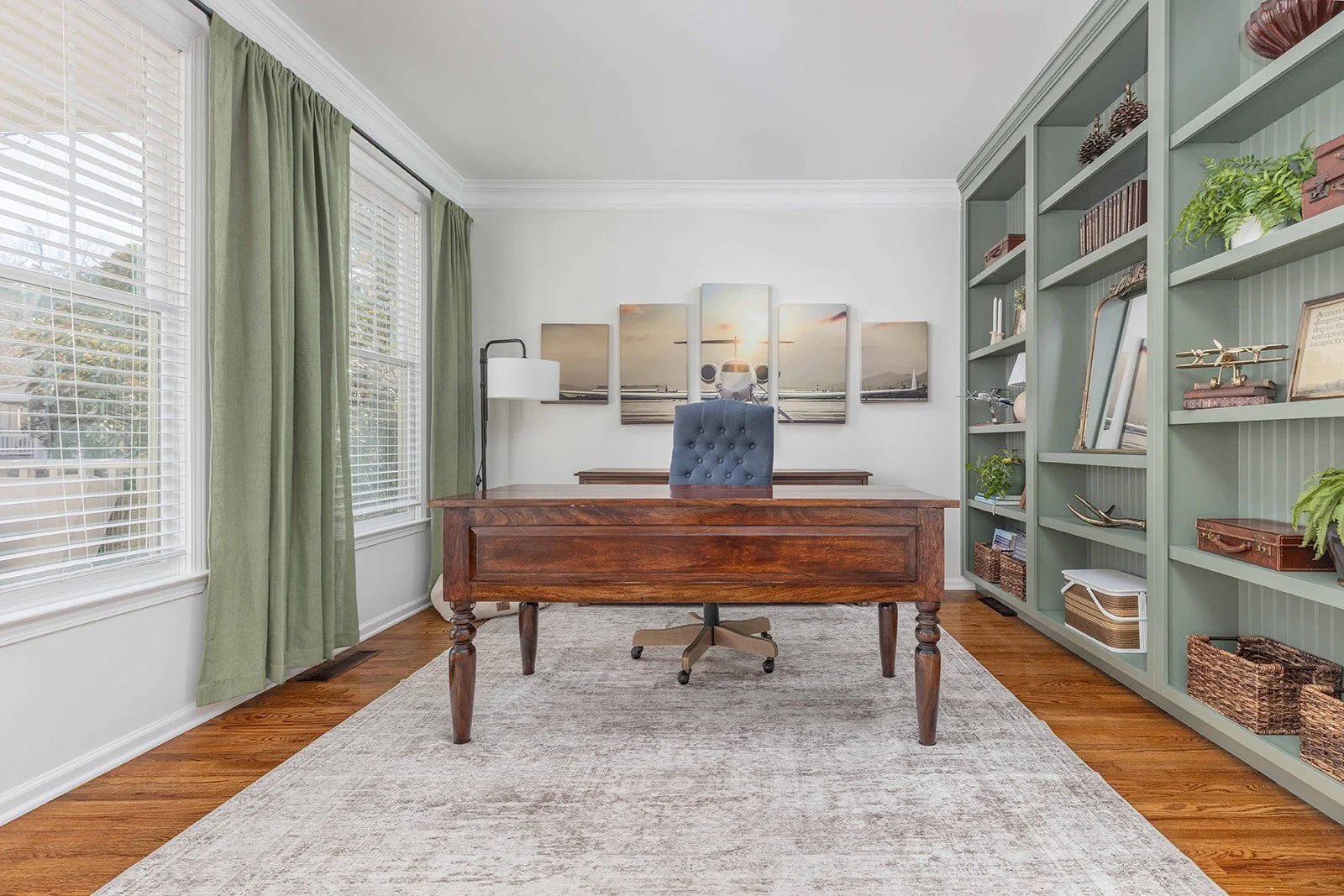 Home office with wooden desk, blue tufted chair, green curtains, large windows, and built-in bookshelf filled with decor items, books, and baskets.