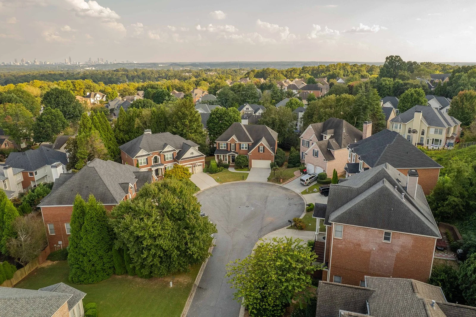 Aerial view of a suburban neighborhood with large houses, green trees, and a circular driveway in the foreground. In the background, there is a city skyline under a partly cloudy sky.