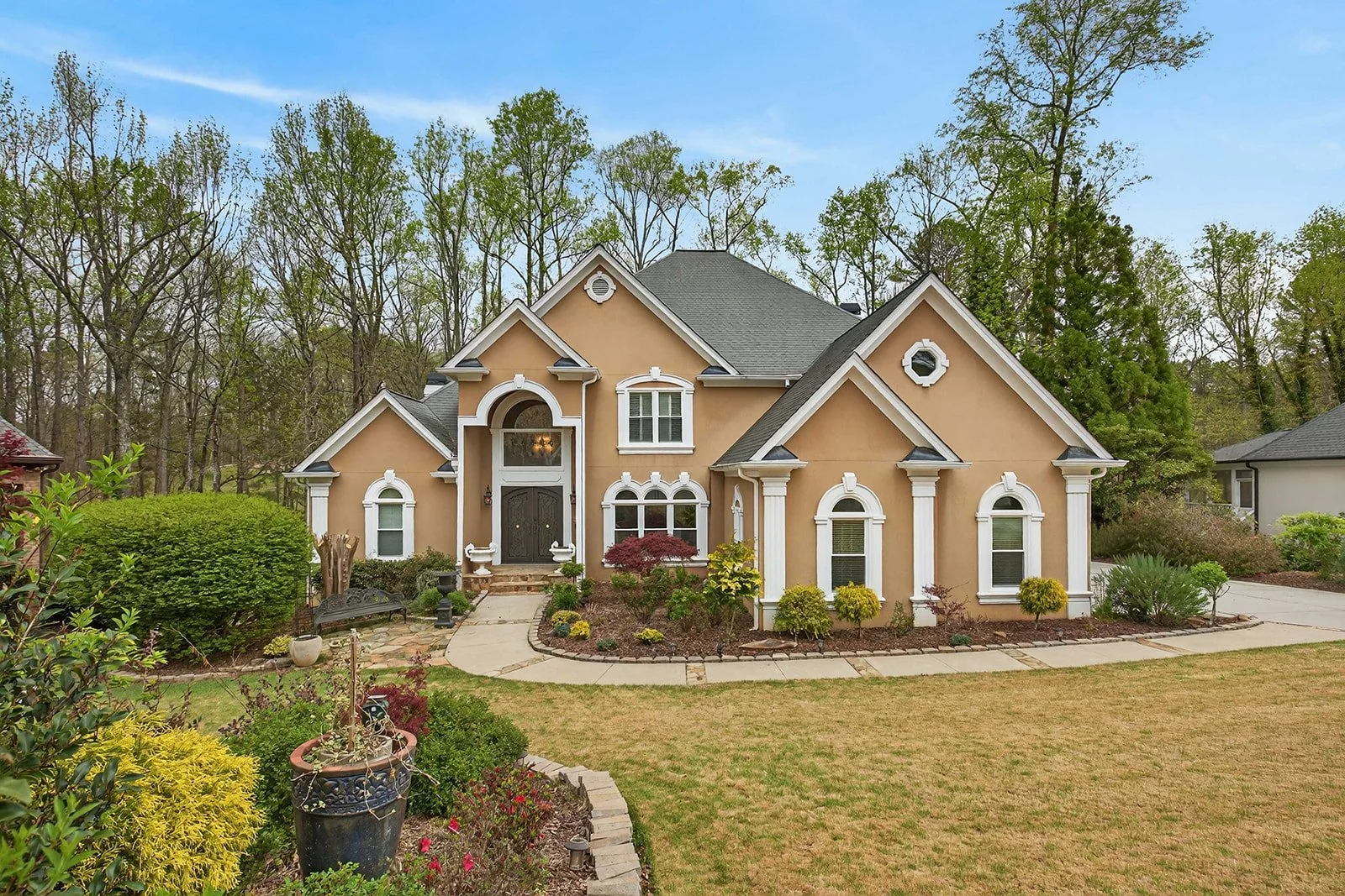 Front view of a two-story beige house with white trim and a gray roof, surrounded by a landscaped garden with various green plants and trees.