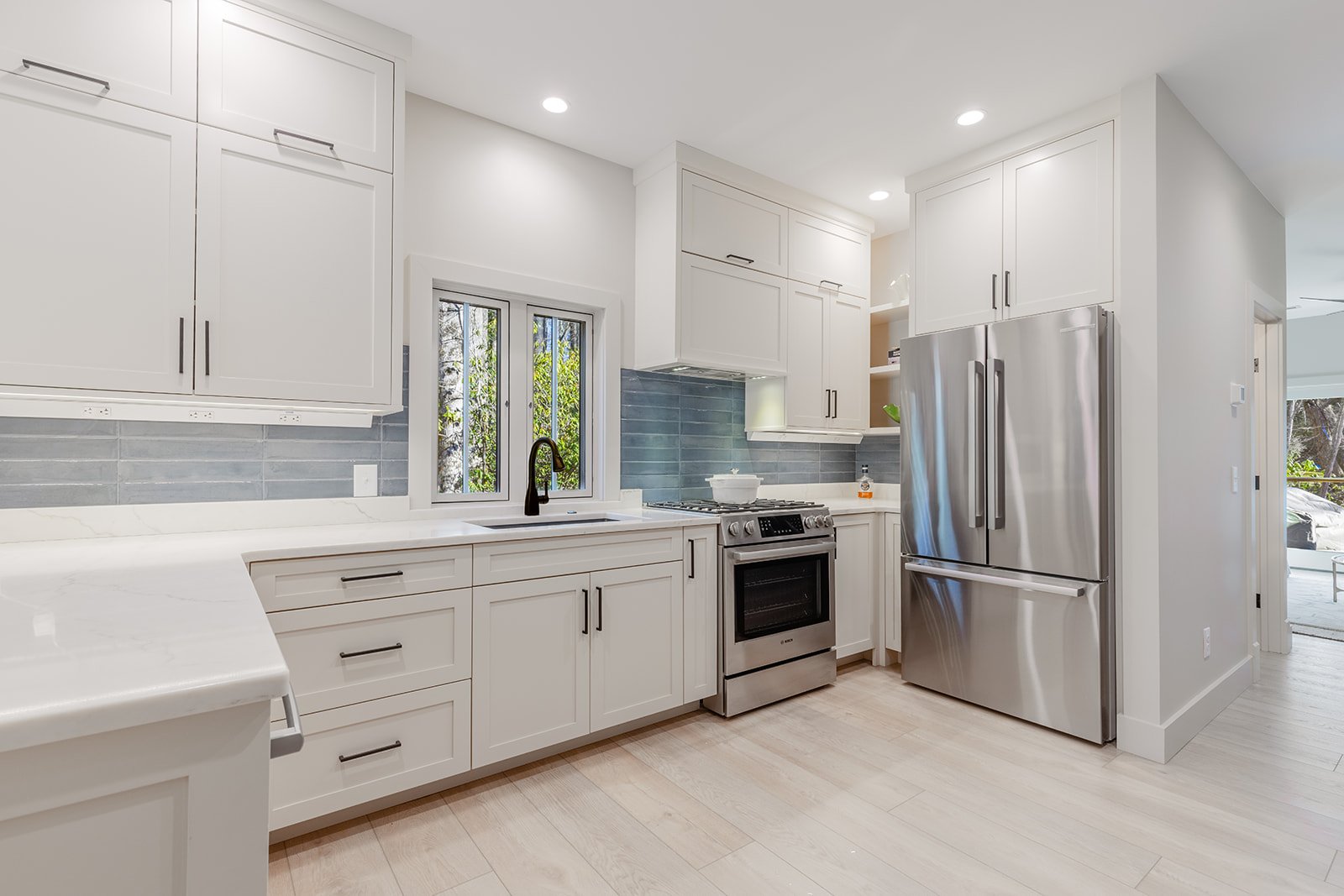 Modern white kitchen with stainless steel refrigerator, gas stove, black faucet, gray backsplash, white cabinets, and light wood flooring.