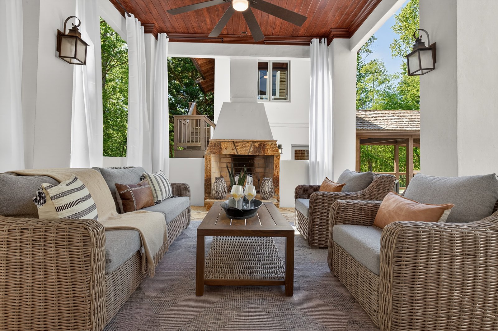 Living room with wicker sofas, throw pillows, a wooden coffee table, a stone fireplace, and large windows with white curtains showing trees outside.