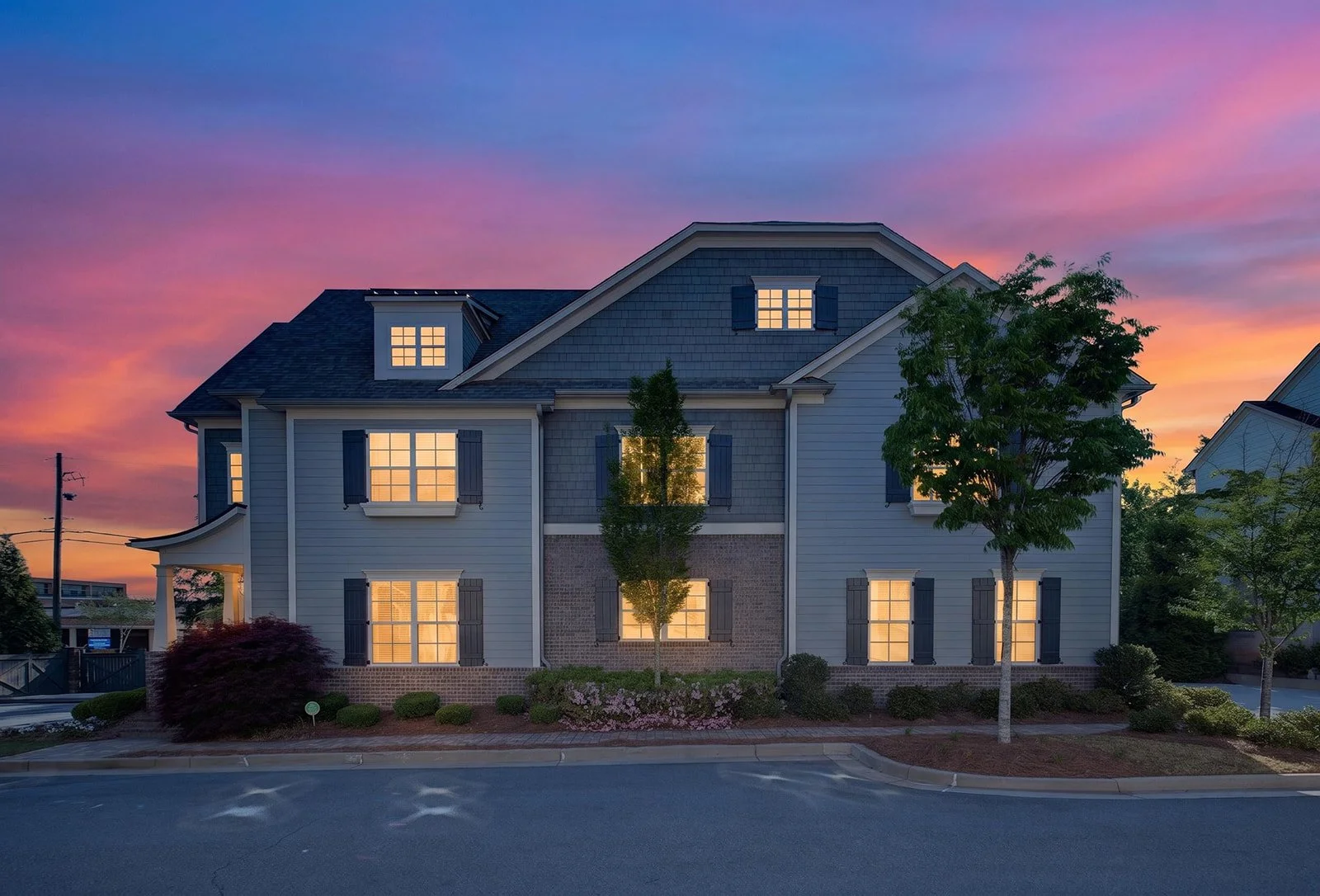 A modern three-story house with lit windows at sunset, surrounded by trees and landscaped bushes.