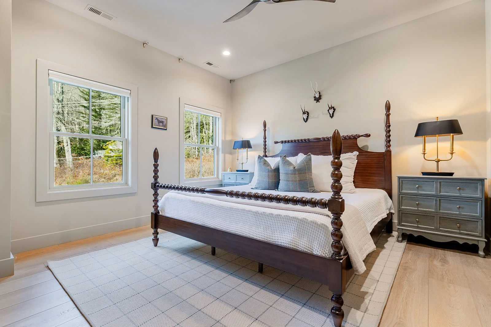 A bedroom with a wooden four-poster bed, two windows showing an outdoor wooded scene, two nightstands with lamps, a beige area rug, and wall decorations including mounted antlers and a framed photo.