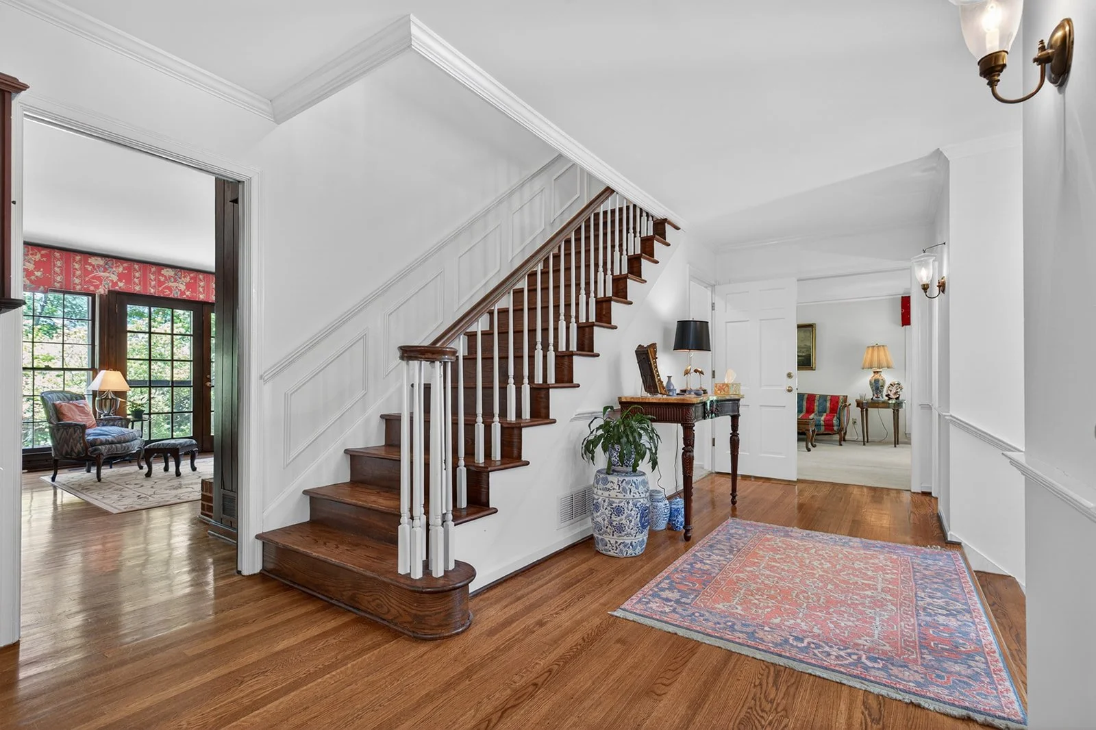 Inside view of a house's entryway with white walls, wooden stairs, an area rug, and various furniture pieces.