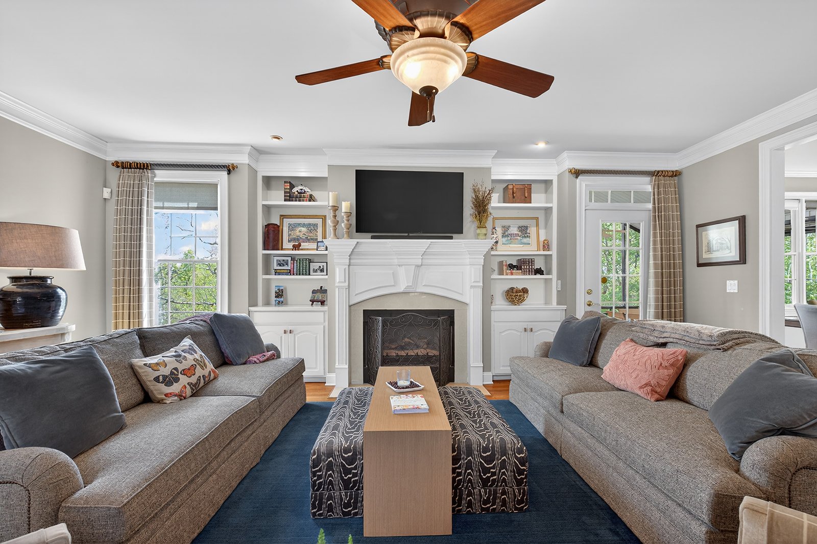 Living room with beige sofas, colorful cushions, a wooden coffee table, a fireplace with a white mantel, built-in shelves on either side, a wall-mounted TV, large windows with checkered curtains, a ceiling fan with wooden blades, and framed artwork.