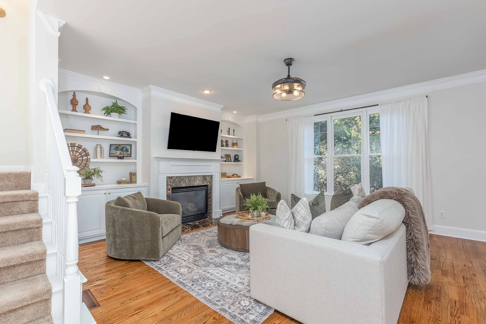 Living room with gray and beige furniture, white walls, wooden floors, a fireplace, built-in shelves, a mounted TV, large windows with white curtains, and a ceiling fan with lights.