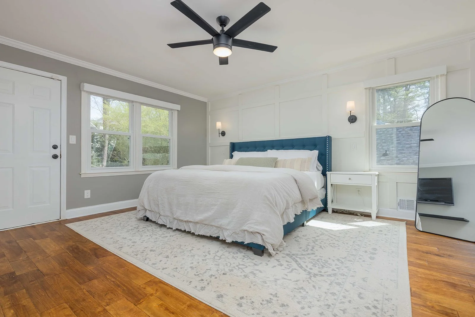 A bedroom with a large bed featuring a blue upholstered headboard, white bedding, and multiple pillows. The room has two windows with white trim, gray walls, a white side table, wall-mounted lamps, a black ceiling fan, a full-length mirror, and hardw