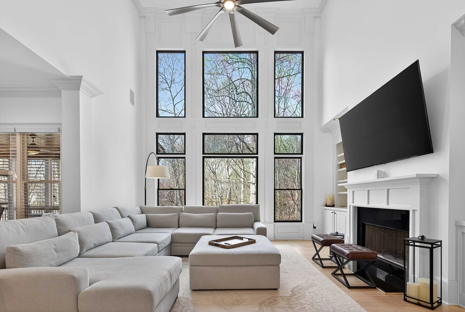 Bright living room with large first-story windows, white walls, cream sectional sofa, fireplace with a black TV above, and a ceiling fan.