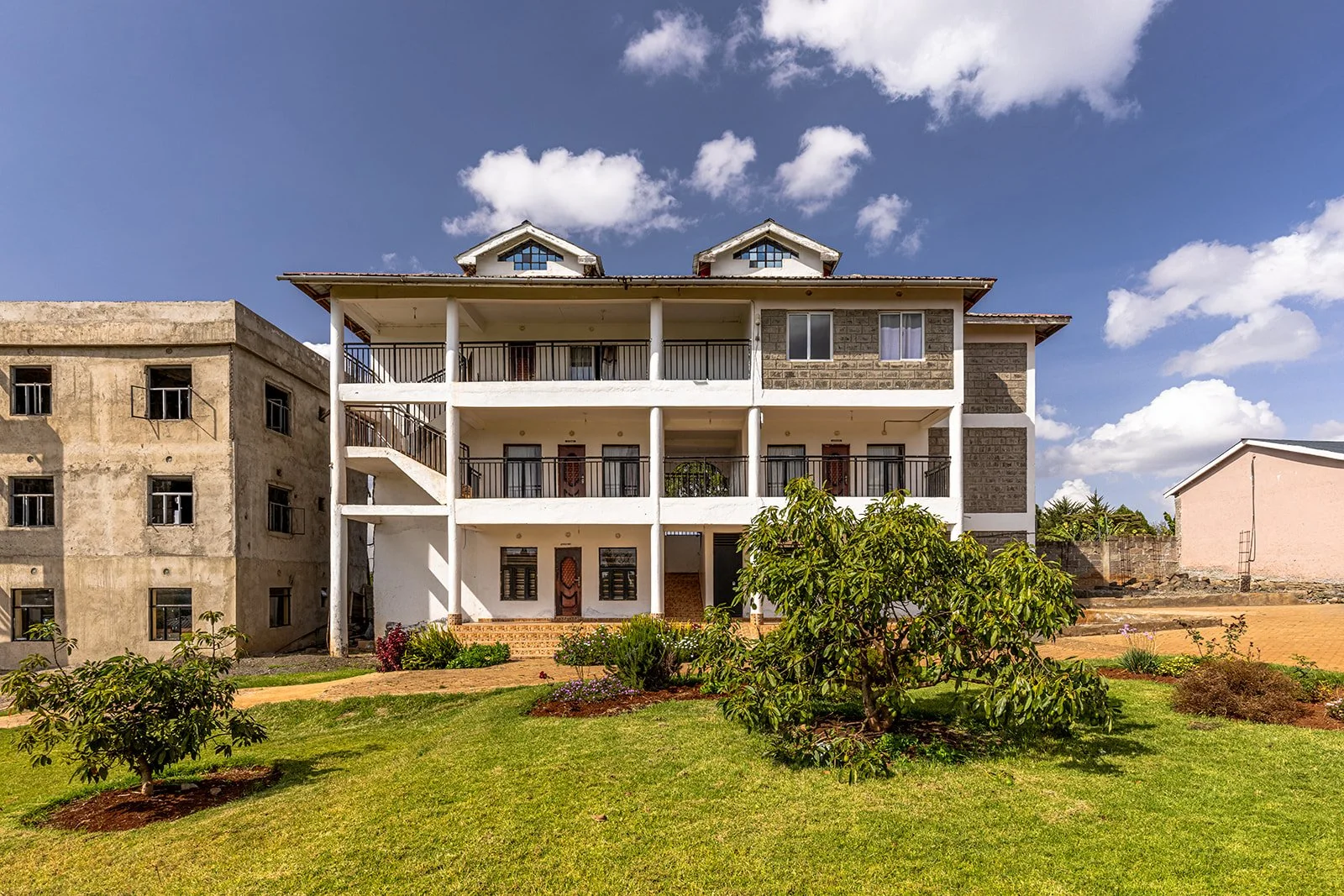 A multi-story residential building with a white facade and black railings, surrounded by a landscaped lawn with small trees and plants, under a partly cloudy sky.
