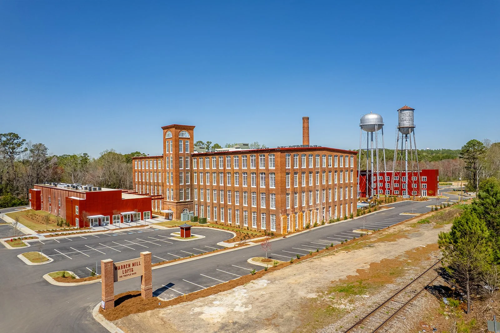 Large brick building with multiple floors and numerous windows, situated near a parking lot, with water towers in the background, and a sign reading "Warren Mill Lofts" at the entrance.