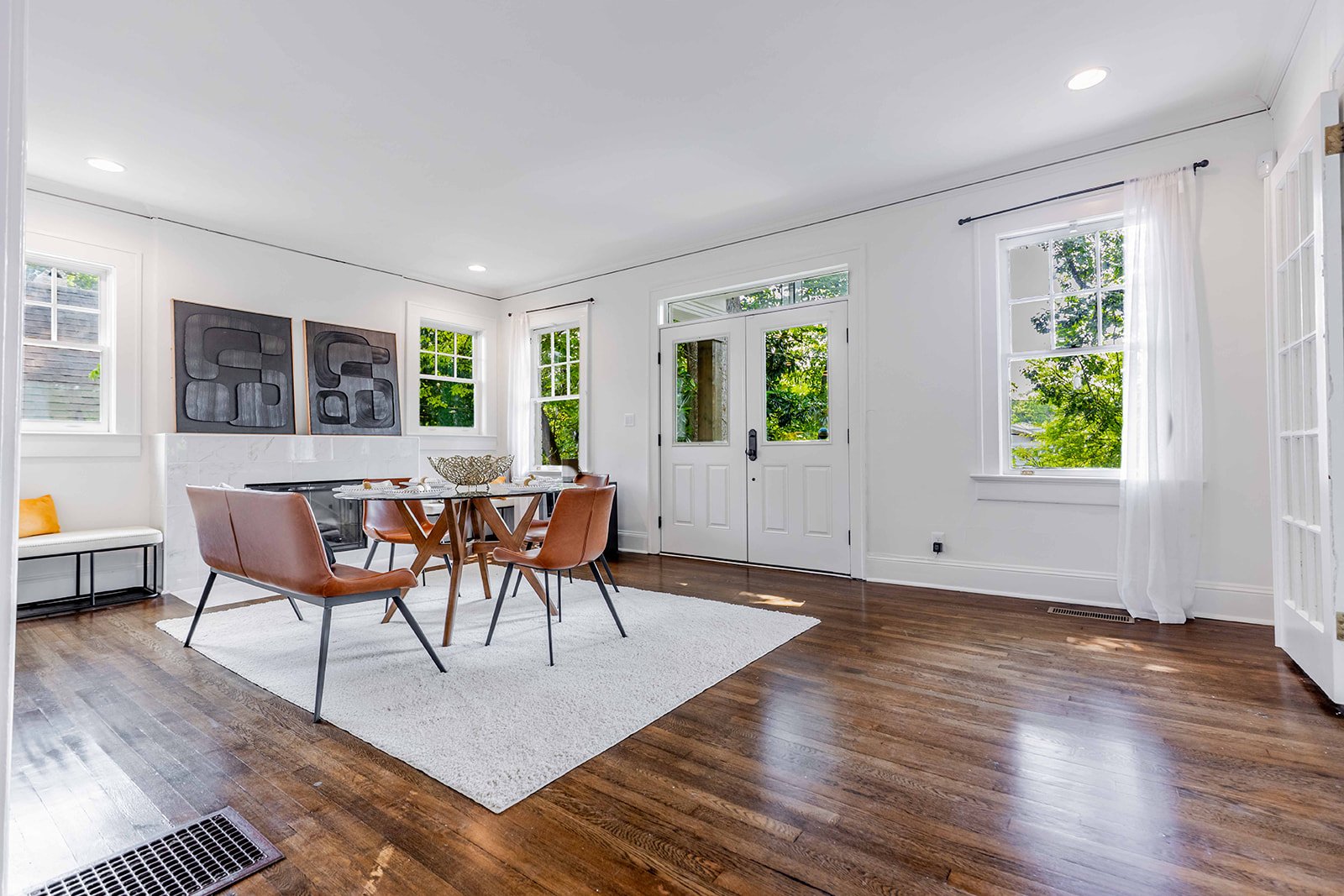 Bright dining room with hardwood floors, white walls, large windows with white curtains, a round wooden table with four brown chairs, abstract wall art, a small bench, and a view of greenery outside.
