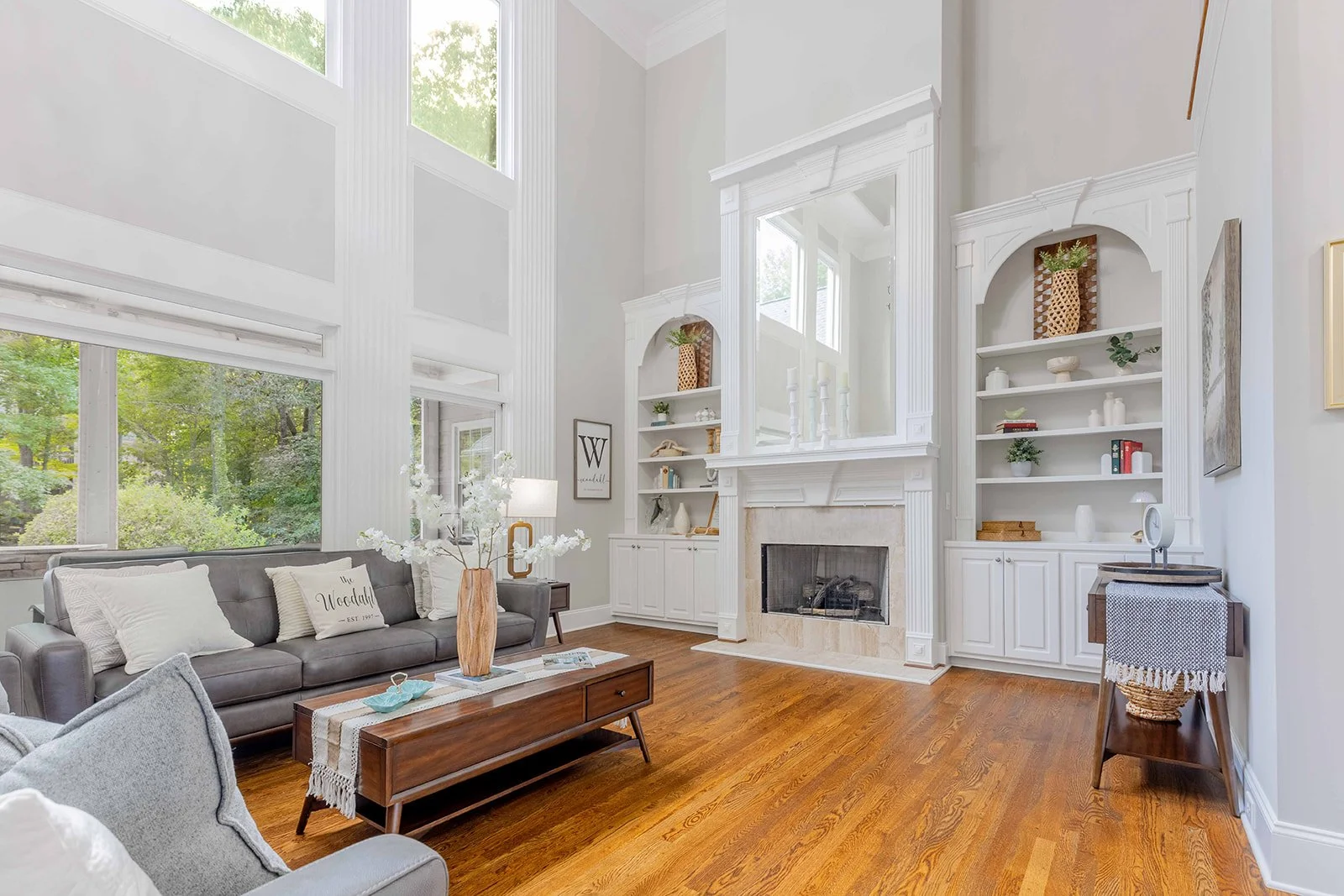Living room with large windows, gray sofa with white pillows, wooden coffee table, built-in white bookshelves around a fireplace, hardwood floors, decorative vases, framed artwork, and a small side table with a basket underneath.