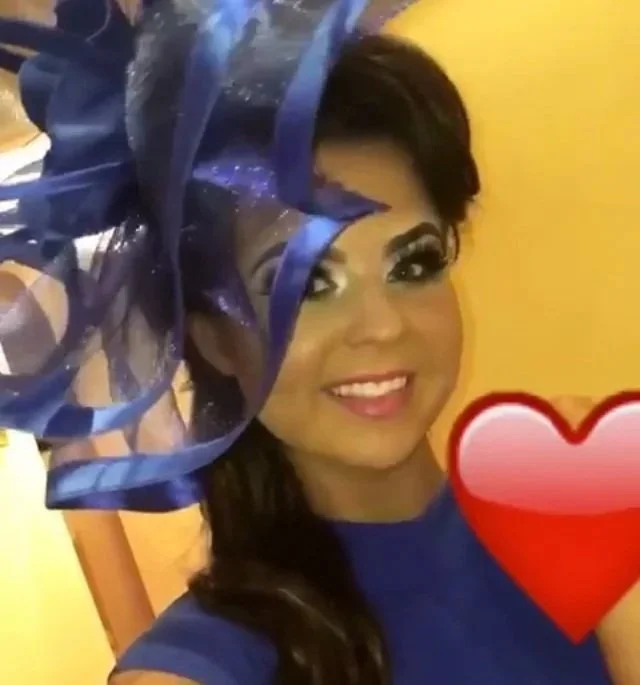 A woman with dark hair, wearing a blue dress and a large decorative blue and purple hat, smiles for a selfie against a yellow background with a red heart emoji overlay.