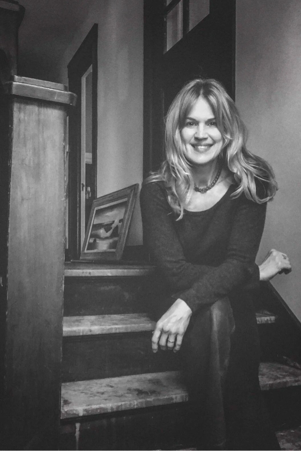 A woman with wavy blonde hair sitting on a wooden staircase, smiling at the camera, dressed in a dark long-sleeve top, with framed pictures on the stairs behind her.
