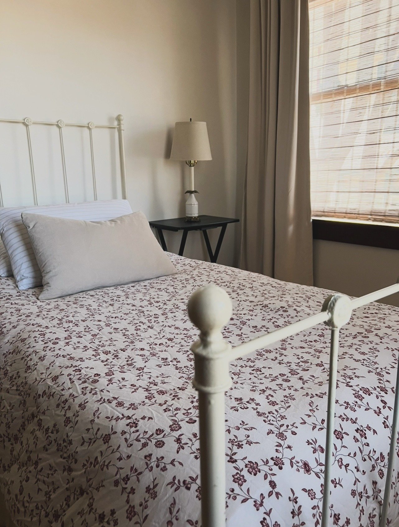 A bedroom with a white metal bed frame with a floral bedspread and pillows, a black side table with a lamp, and a window with blinds and curtains.