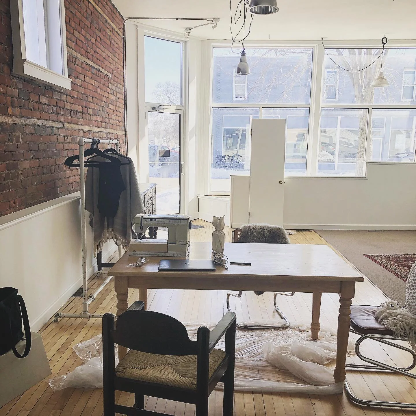 Bright room with large windows, brick wall, and hardwood floor. Furniture includes a wooden table with sewing machine, chairs, a folding chair with fur, and a chair with a blanket. Items include a paper bag, a notebook, and some plastic wrapping. There is a clothing rack with clothes, and ceiling wires hanging down. Outside the windows, a street with parked bicycles and houses is visible.
