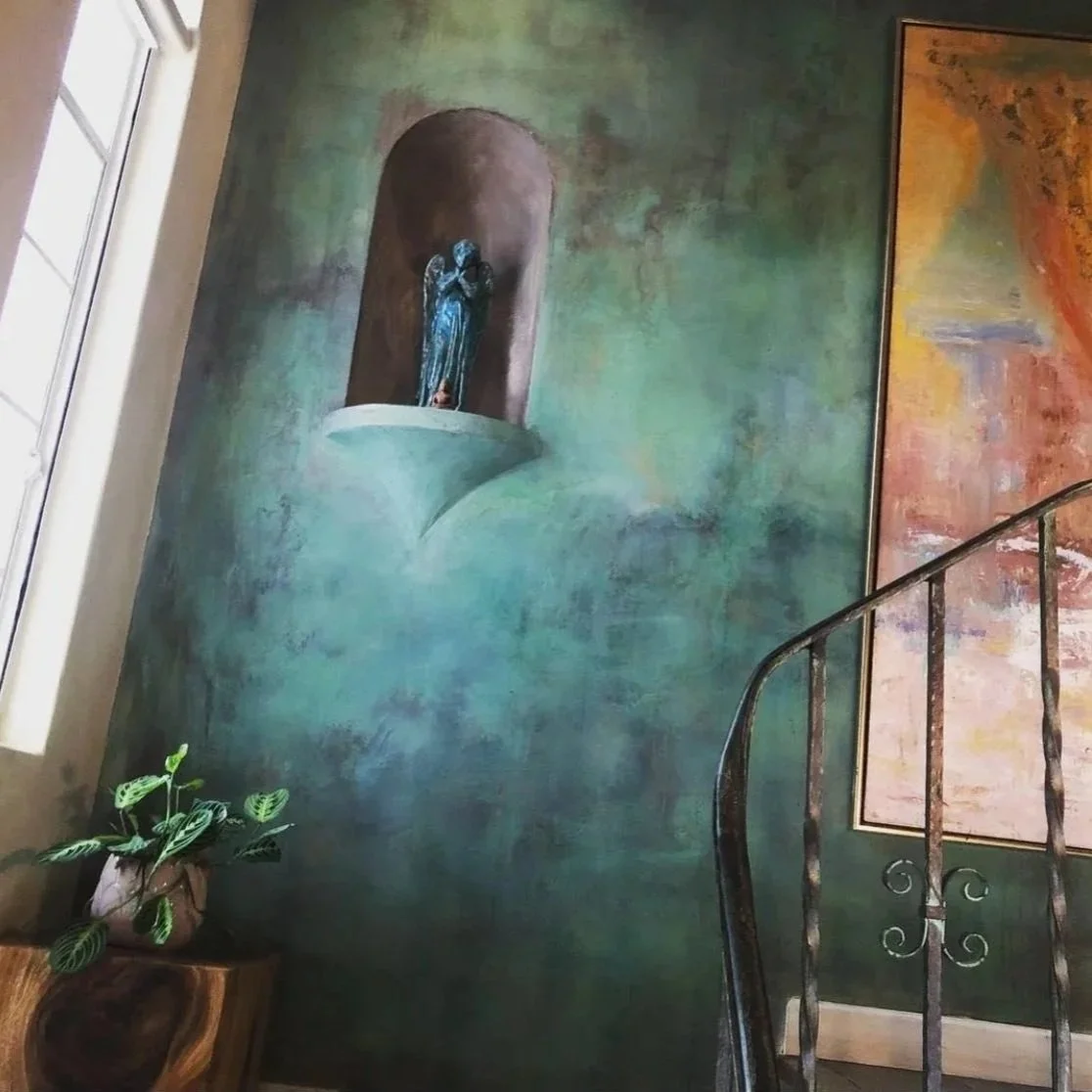 Interior of a room with a large mural on the wall depicting a faceless figure with a small statue in a niche where the face should be. There is a plant in a pot on a wooden side table and part of a metal railing staircase is visible.