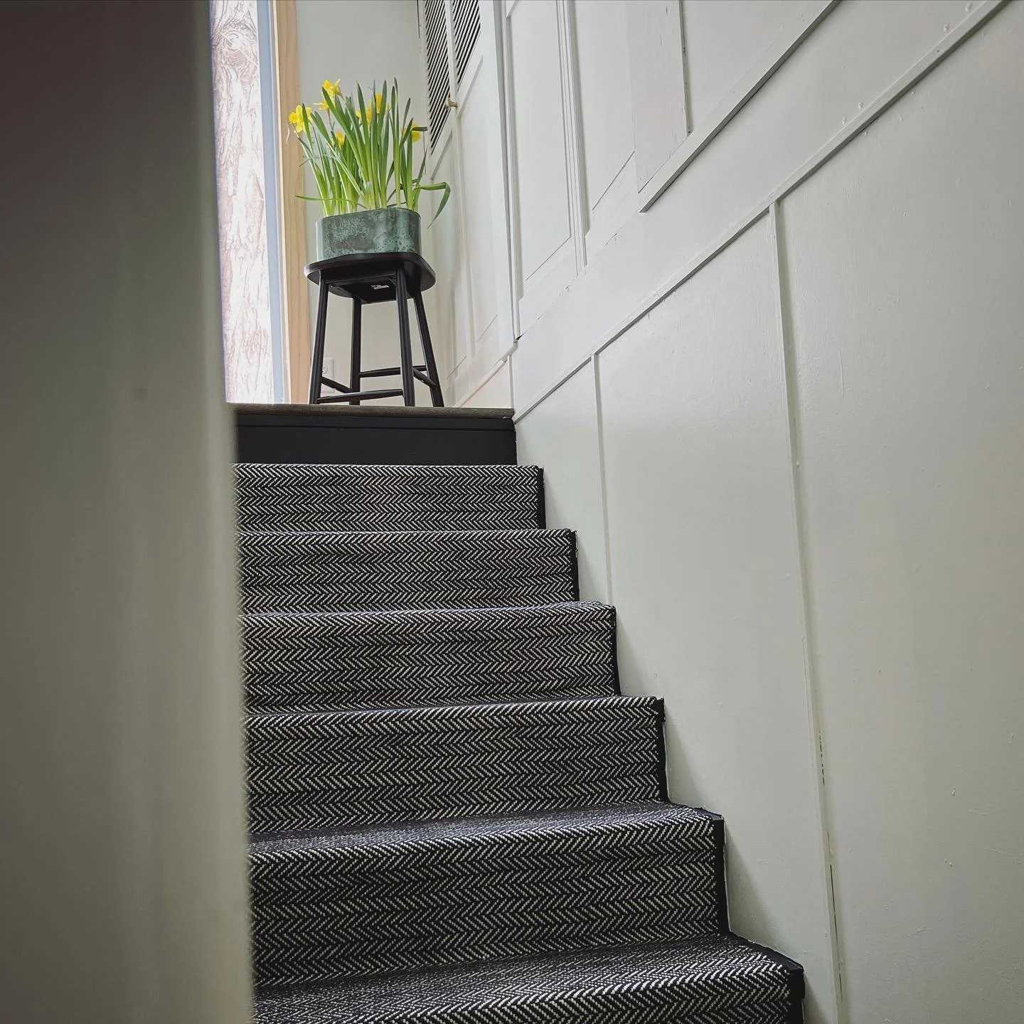A staircase with patterned carpet leading up to a landing, on which a black stool holds a green potted plant with yellow flowers, near a window with wooden frame.