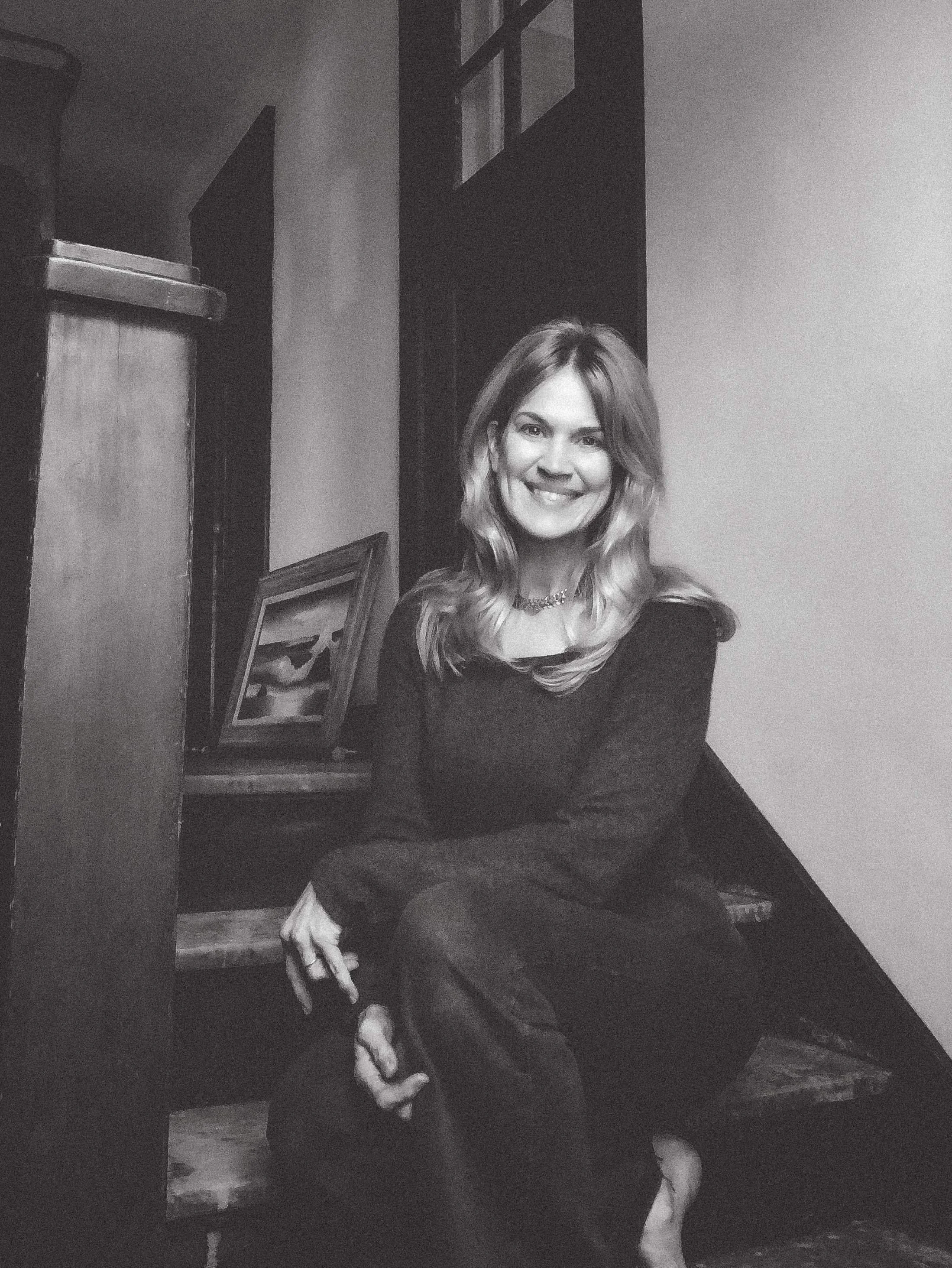 A woman with long wavy hair wearing a dark dress, smiling, sitting on a wooden floor with a framed picture leaning against the wall behind her.