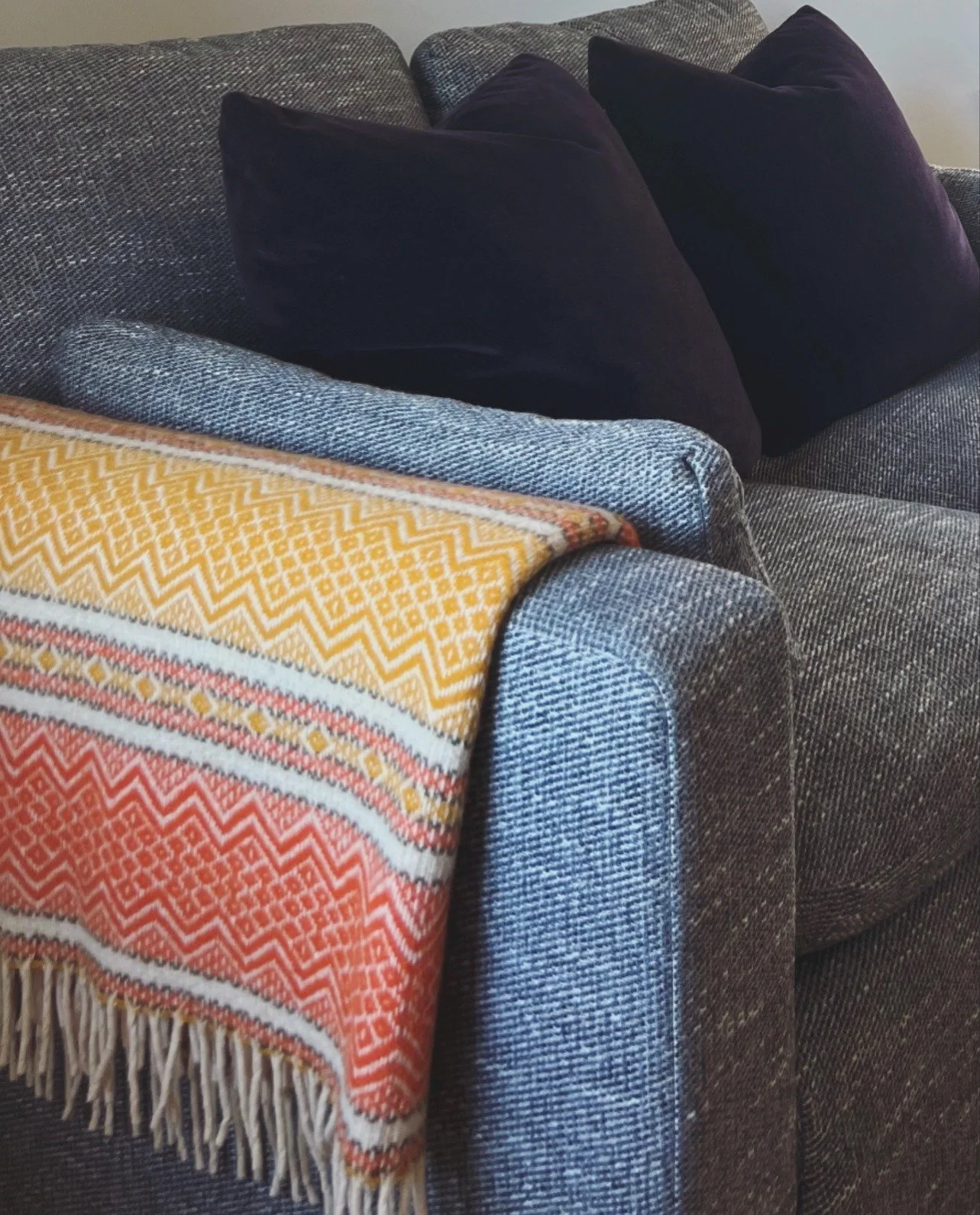 A gray upholstered couch with two black pillows and a colorful patterned blanket with fringes draped over the armrest.