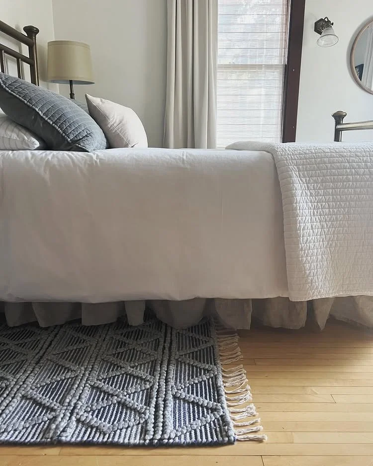 A neatly made bed with white bedding, gray pillows, and a quilted white blanket in a bright bedroom with a window, curtains, a round mirror, a wall-mounted light, a nightstand with a lamp, a patterned rug, and hardwood floors.