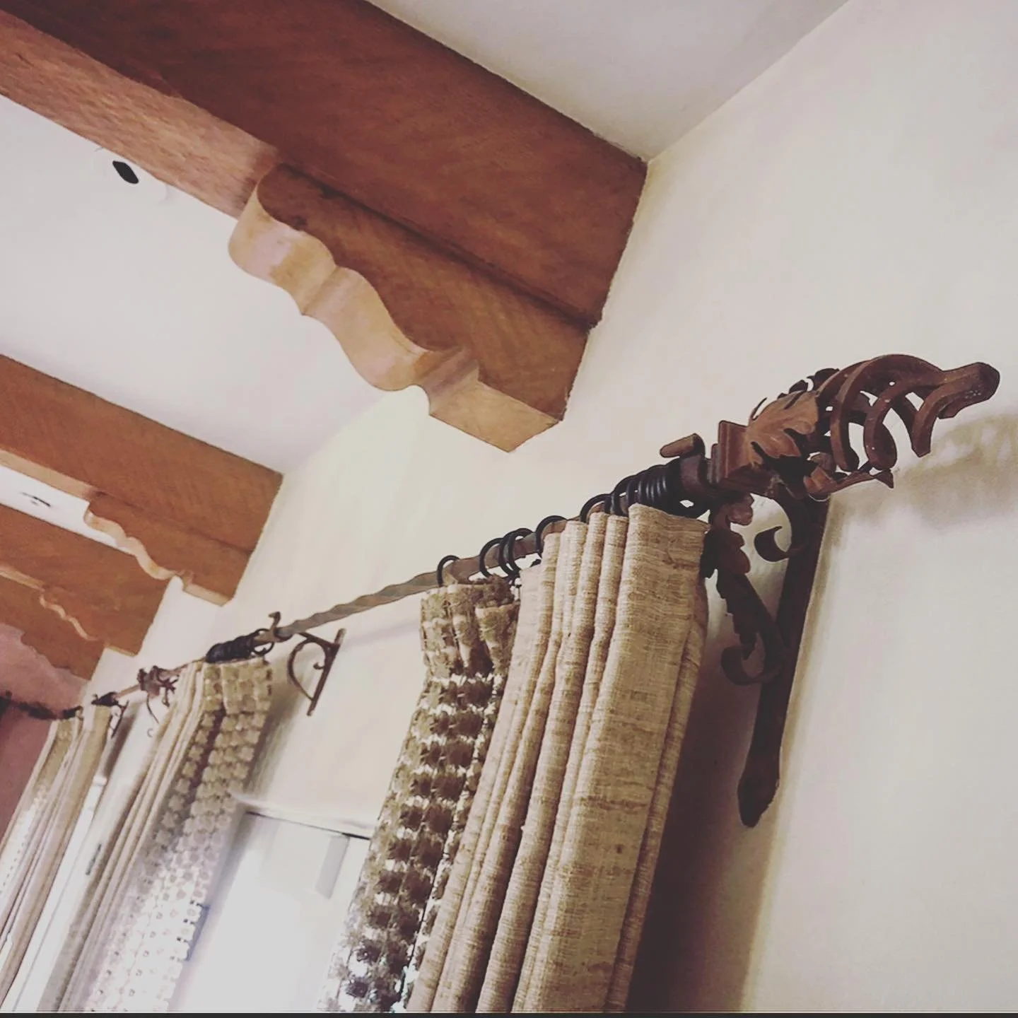 A room corner with beige curtains on a black wrought iron curtain rod, and wood beams on a white ceiling.