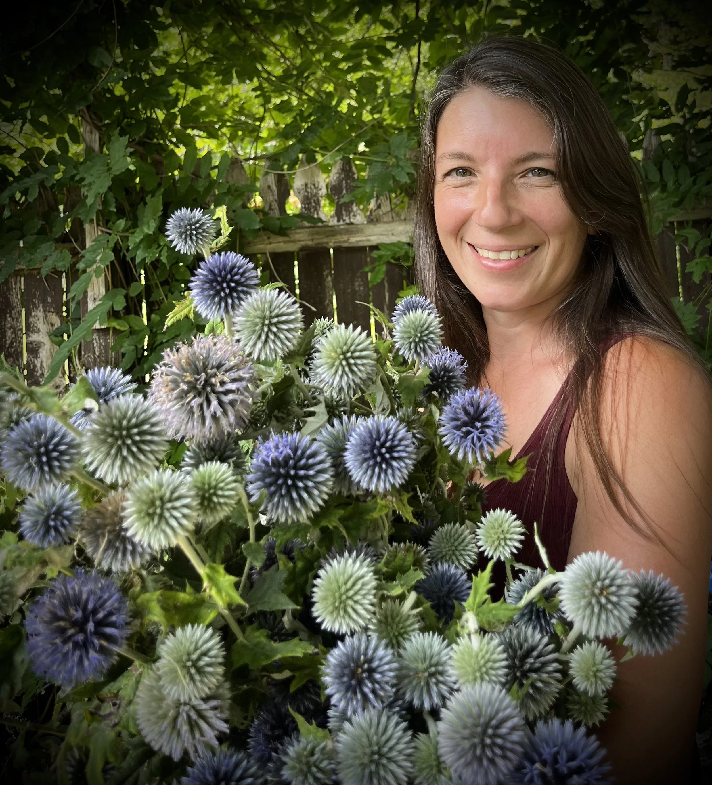 Smiling woman with long brown hair poses outdoors with a large bouquet of blue and white spiky flowers, surrounded by green foliage and a wooden fence in the background.