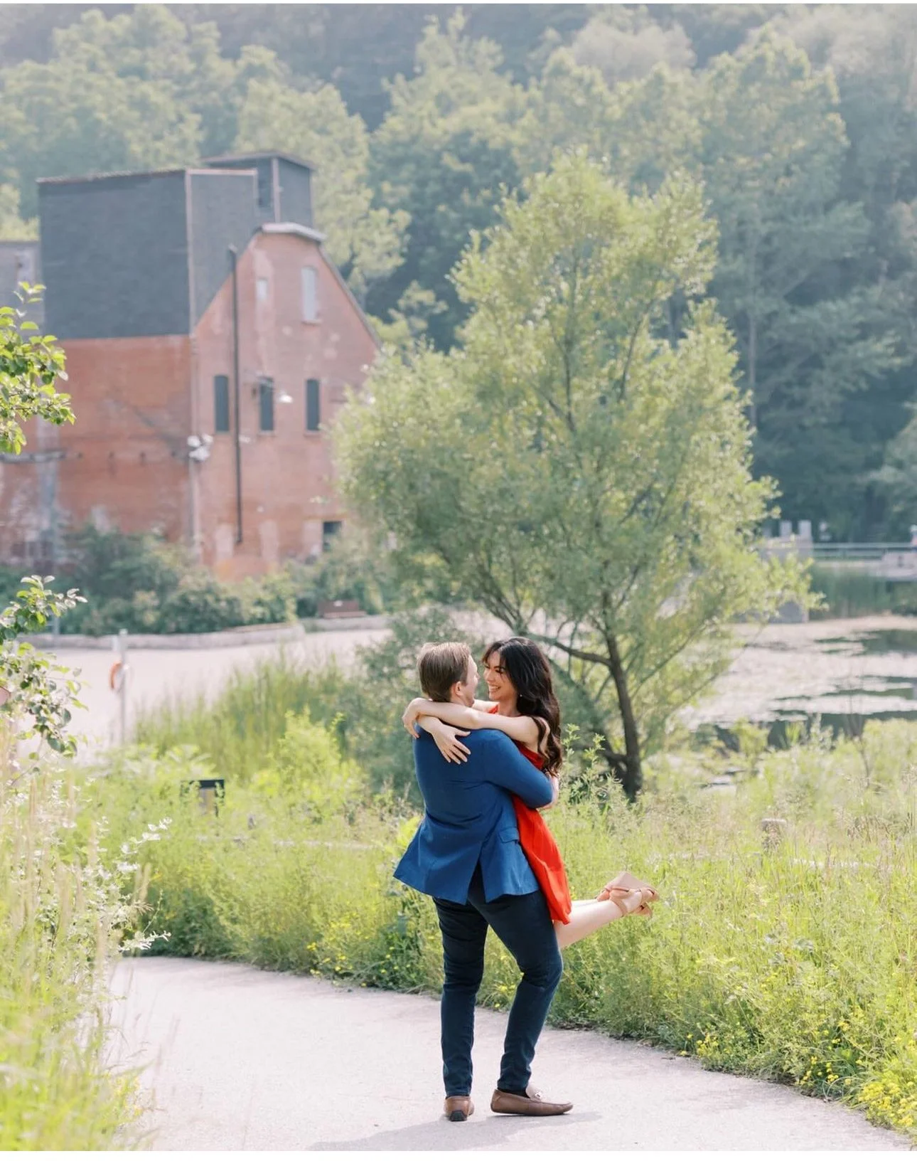 A man and woman embrace on a nature trail, smiling at each other. The man lifts the woman in his arms, her arms around his neck. They are in a green, grassy area with trees and a river in the background.