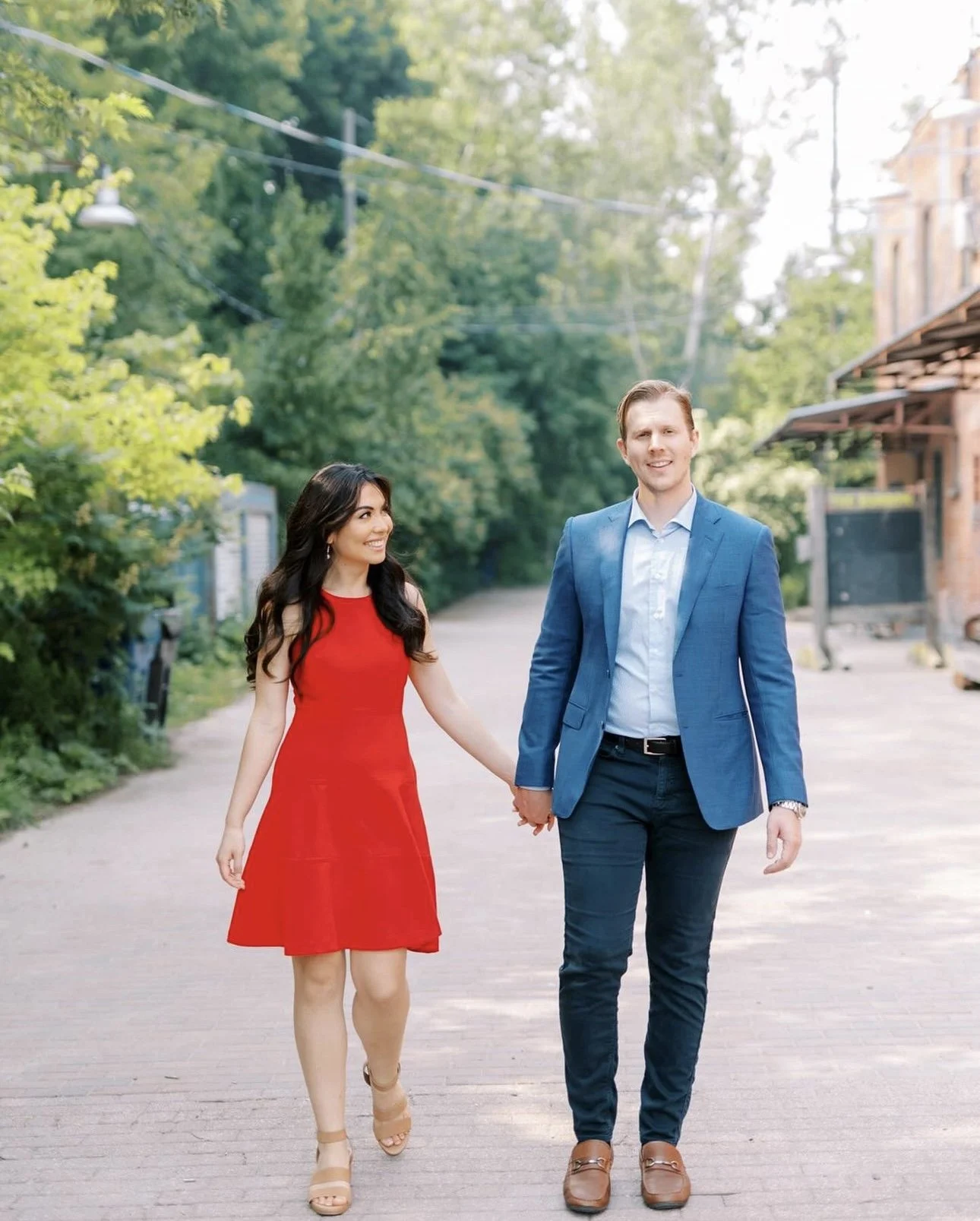A couple walking hand in hand outdoors on a sunny day, surrounded by greenery. The woman is wearing a red dress and beige heels, and the man is dressed in a blue blazer, white shirt, dark pants, and brown shoes.