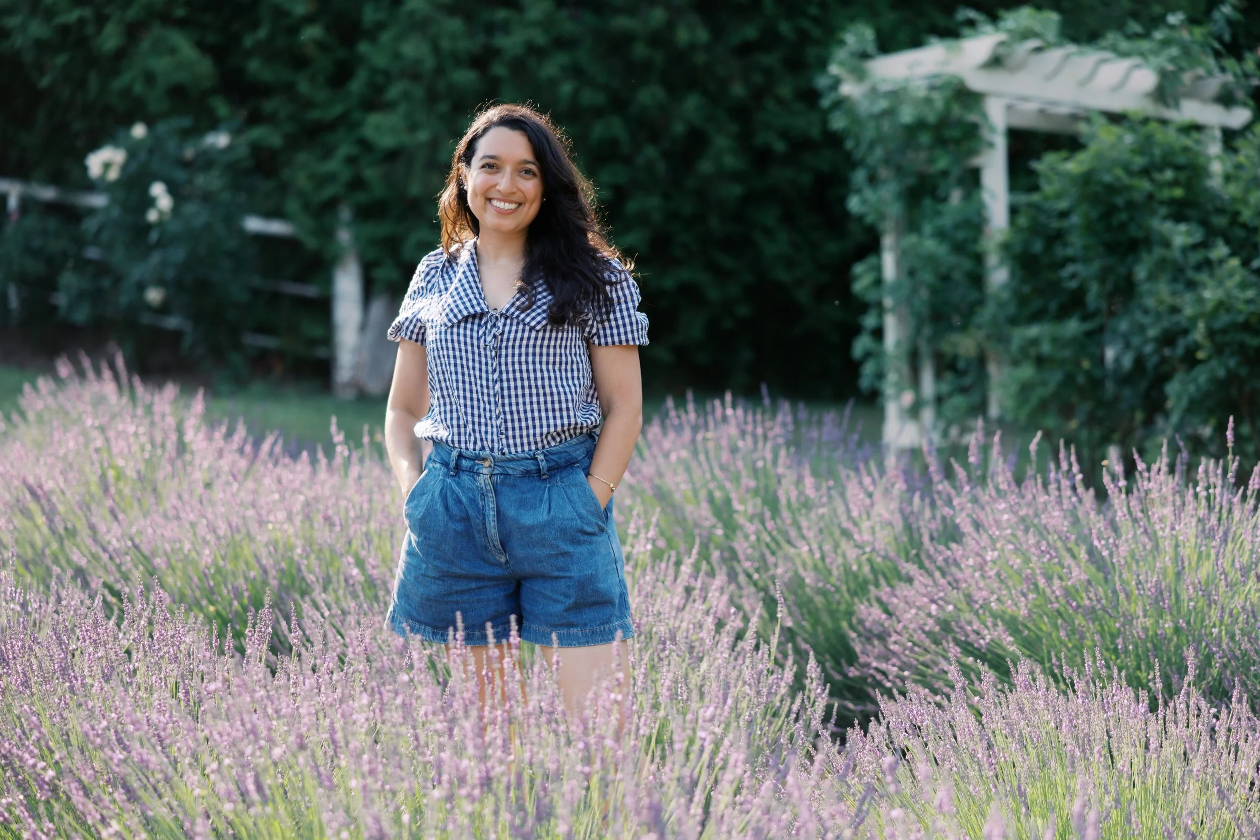 A woman with long dark hair wearing a blue and white checkered shirt and denim shorts standing in a field of purple flowers with green trees in the background.