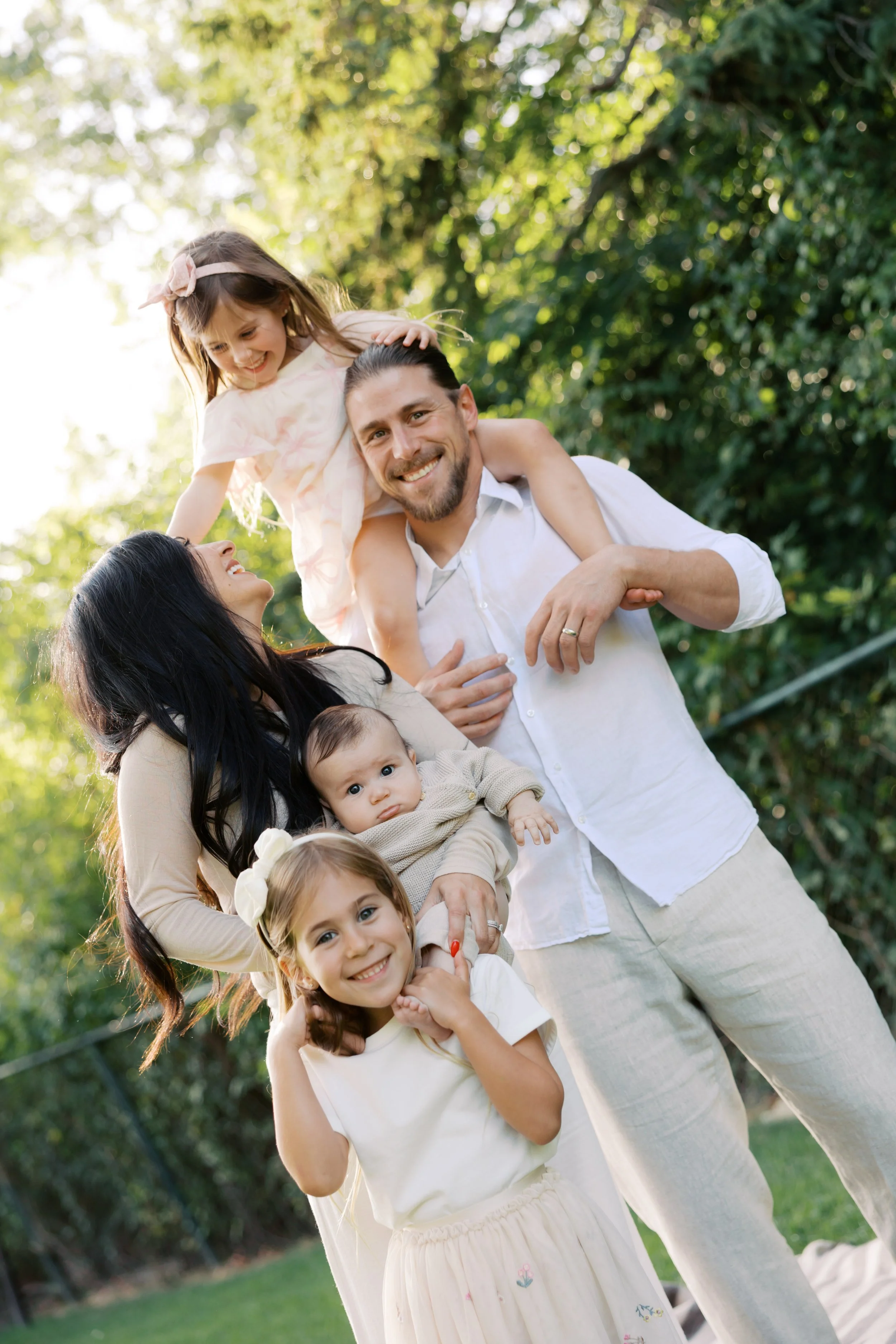 A family of six enjoying time outdoors in a grassy area with trees in the background. The adults, a man and a woman, are smiling and holding their children. The children, three girls and one baby, are smiling and looking at the camera. The girl on th
