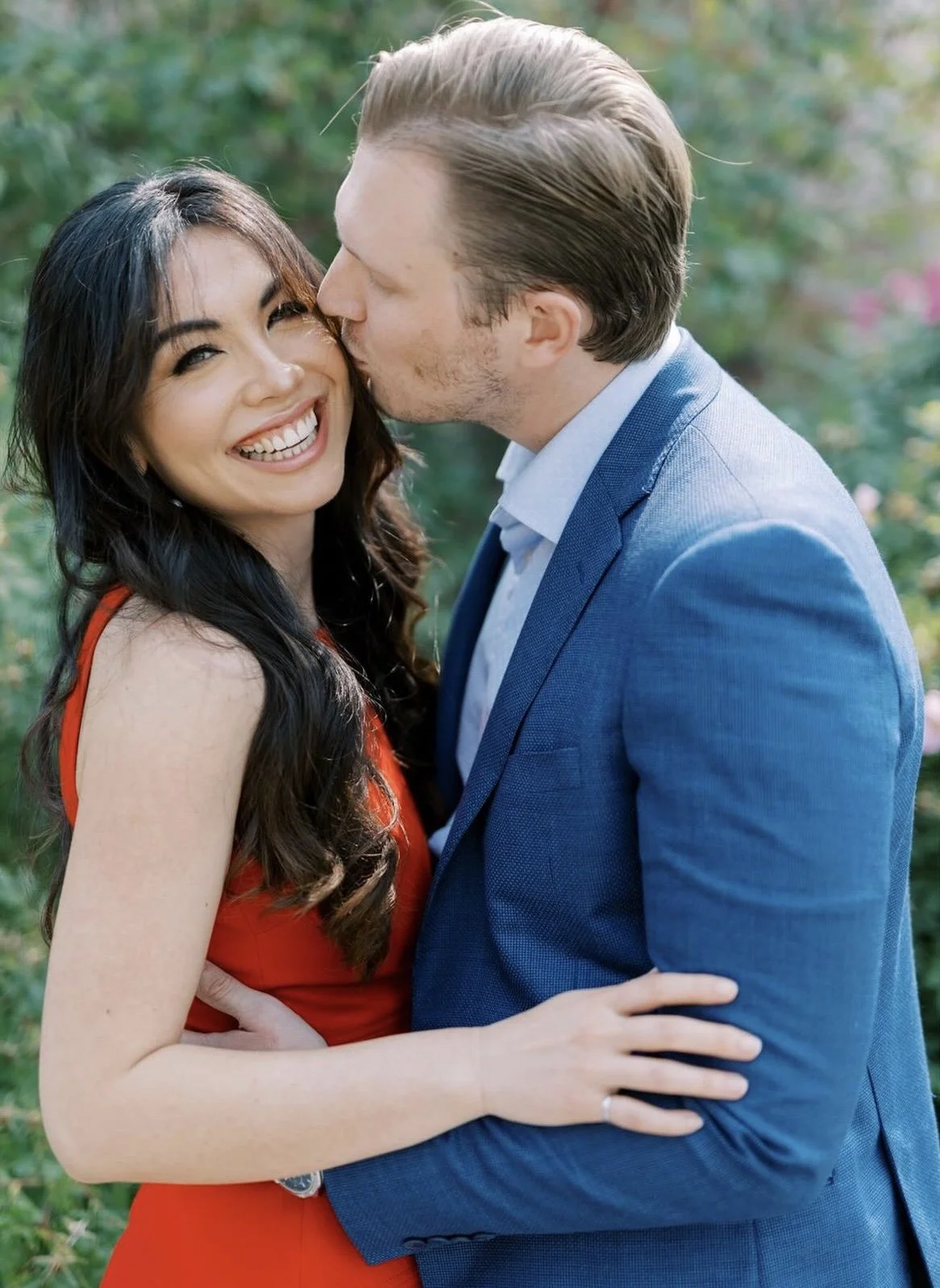 Happy couple, woman in red dress smiling and man in blue suit, kissing outdoors in garden.