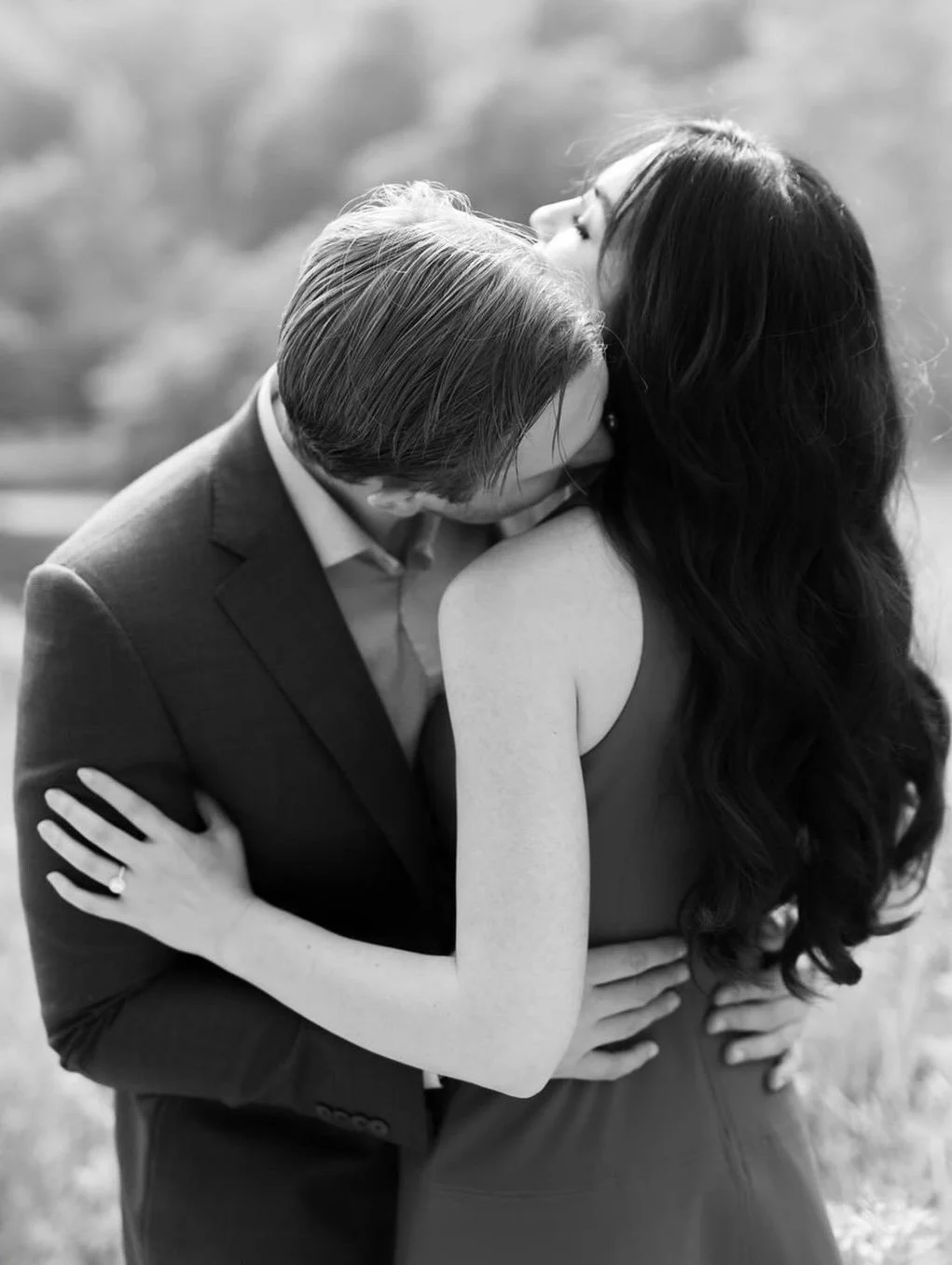 A black-and-white photograph of a man and woman embracing outdoors. The man is in a suit, leaning in for a kiss, with his face close to the woman's shoulder. The woman has long dark hair and is wearing a sleeveless dress; her eyes are closed as she t