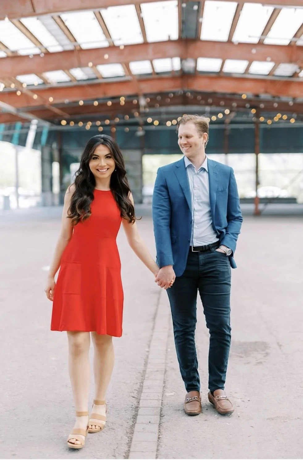 A smiling woman in a red dress holding hands with a man in a blue blazer and dark jeans, walking inside a covered outdoor area with string lights overhead.