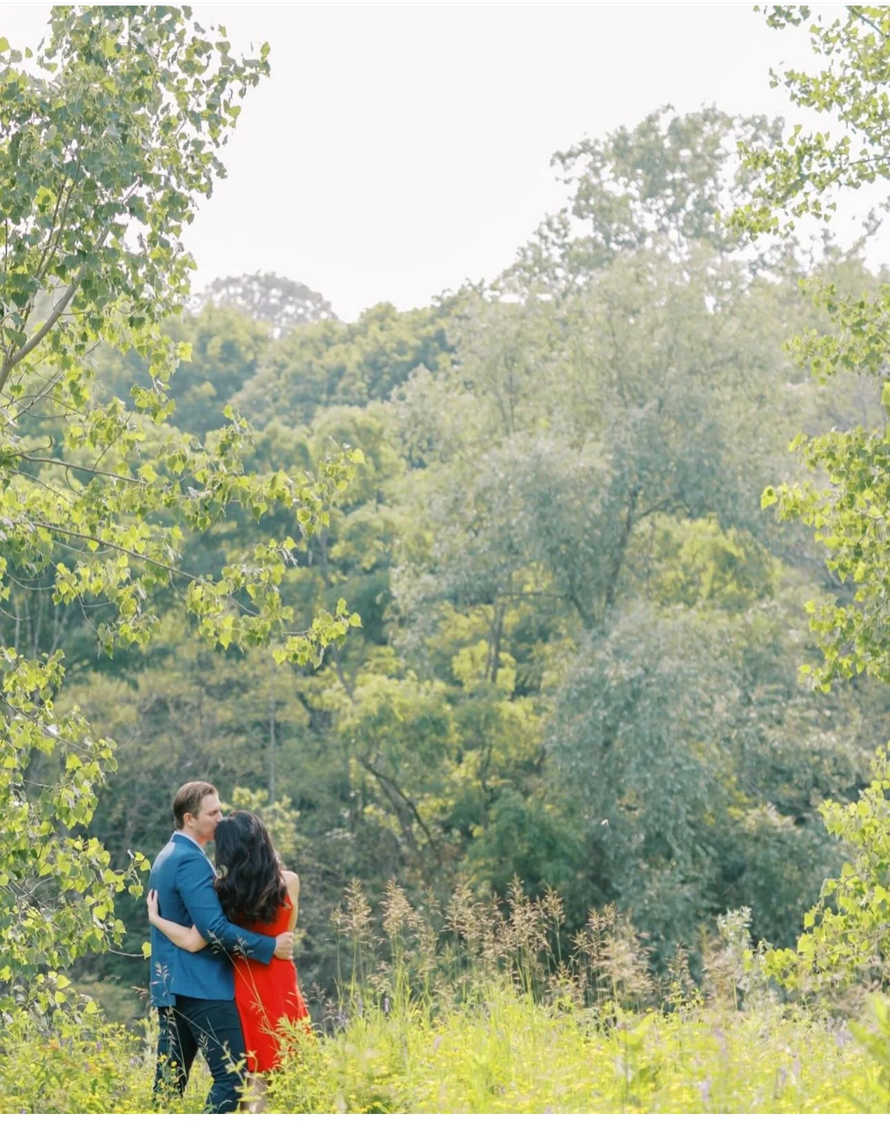 A couple hugging in a grassy field with trees and a forested hill in the background.